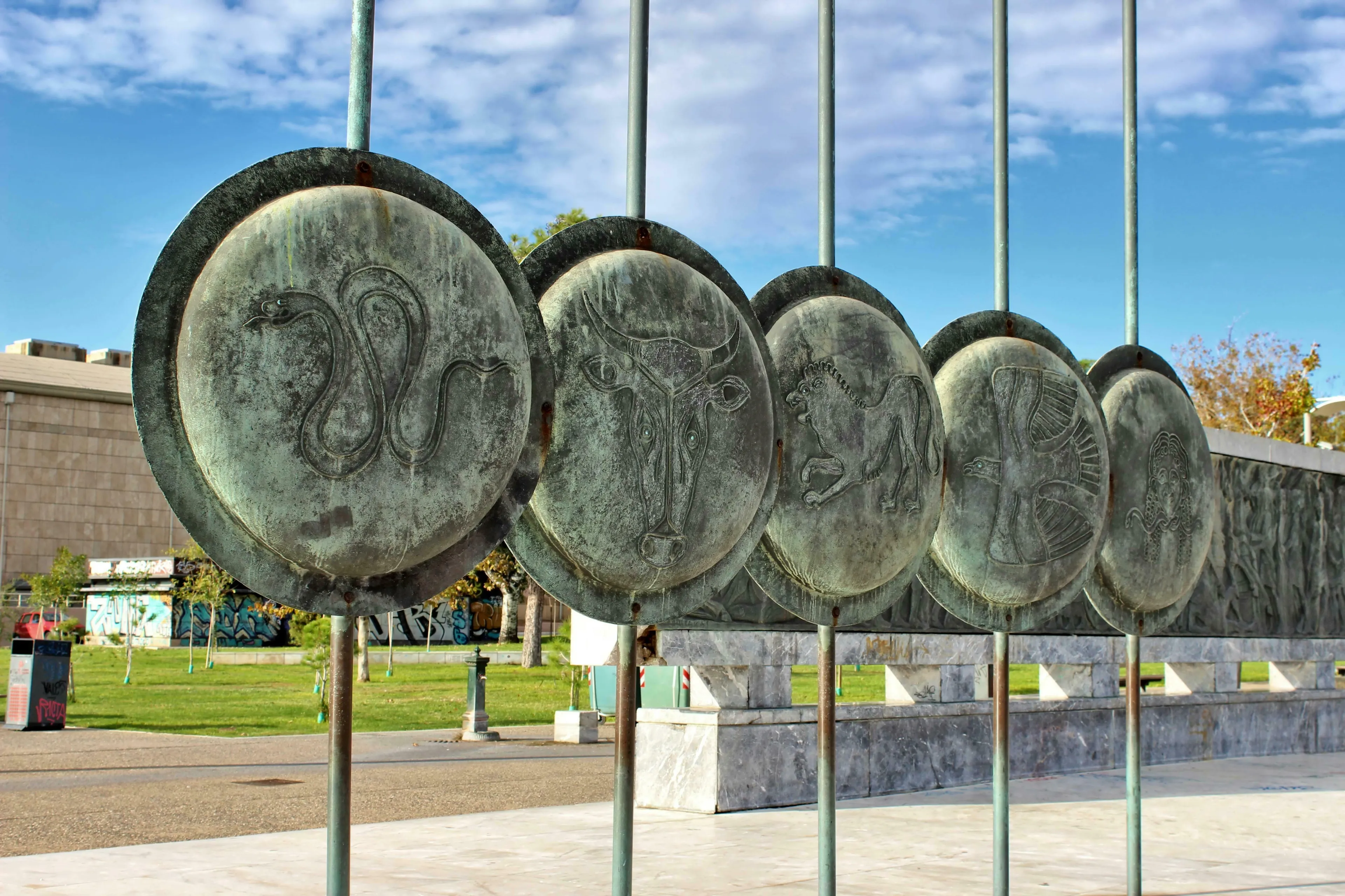Bronze shields with animal engravings from Greek Mythology, resembling the Chimera, line a sunny outdoor path.