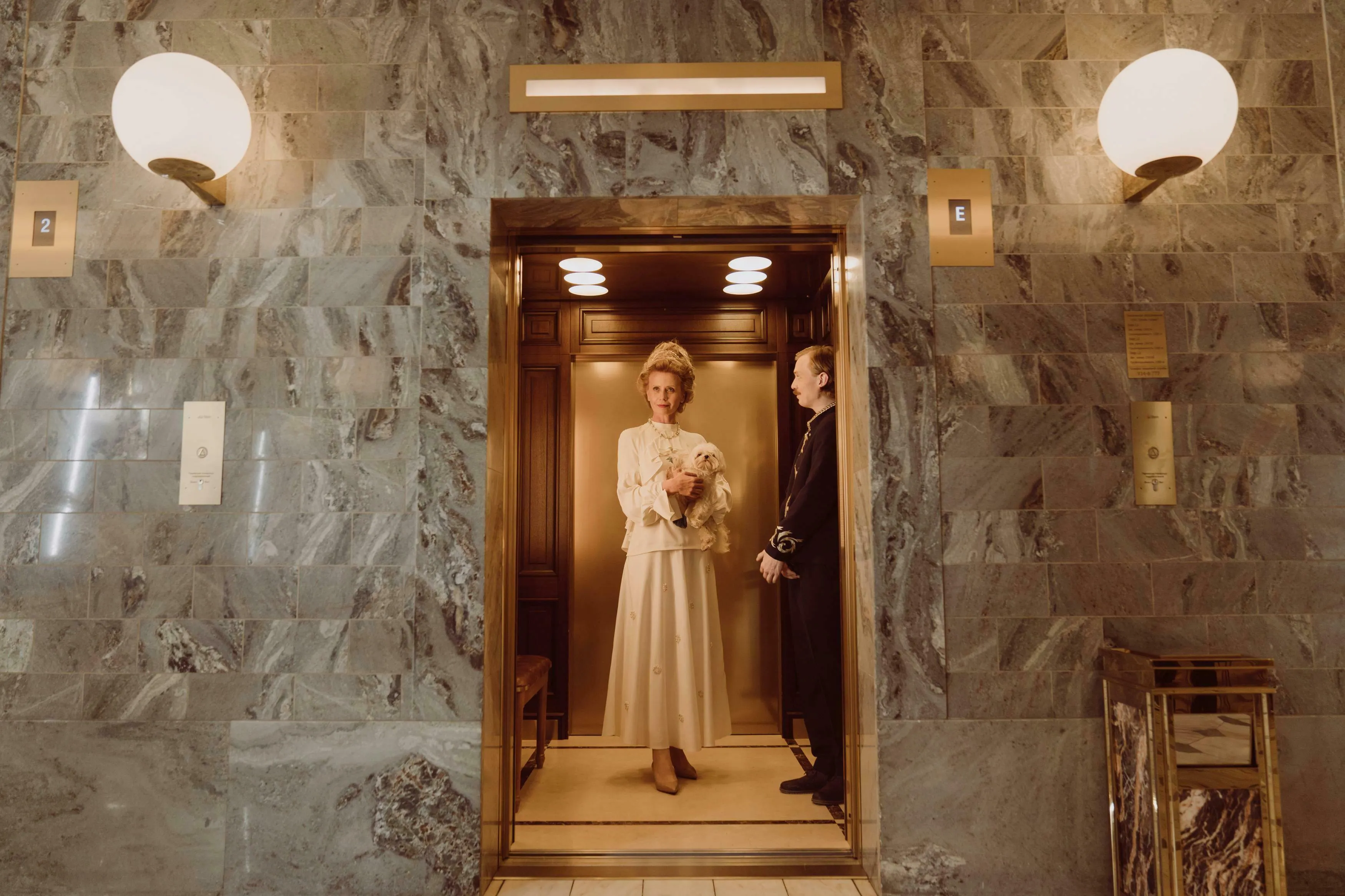 Woman holding a dog and a bellboy standing in a London hotel elevator, surrounded by marble walls. Great British Vocabulary Quiz.