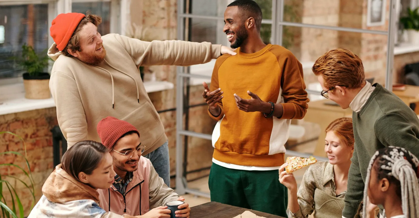 A group of diverse friends laughing and talking, with one eating pizza. They might be discussing The Great British Vocabulary Quiz.