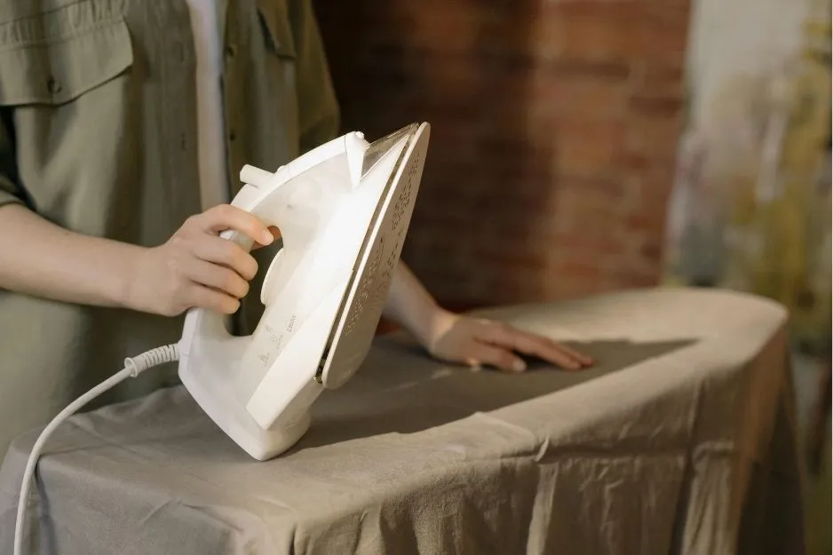 Close-up of a person's hands holding a white iron, positioned over a grey ironing board, performing ironing clothes.