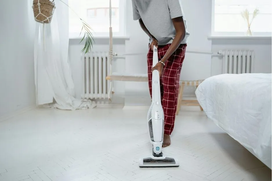 A person in plaid pajamas is vacuuming the white wooden floor of a sunlit bedroom.