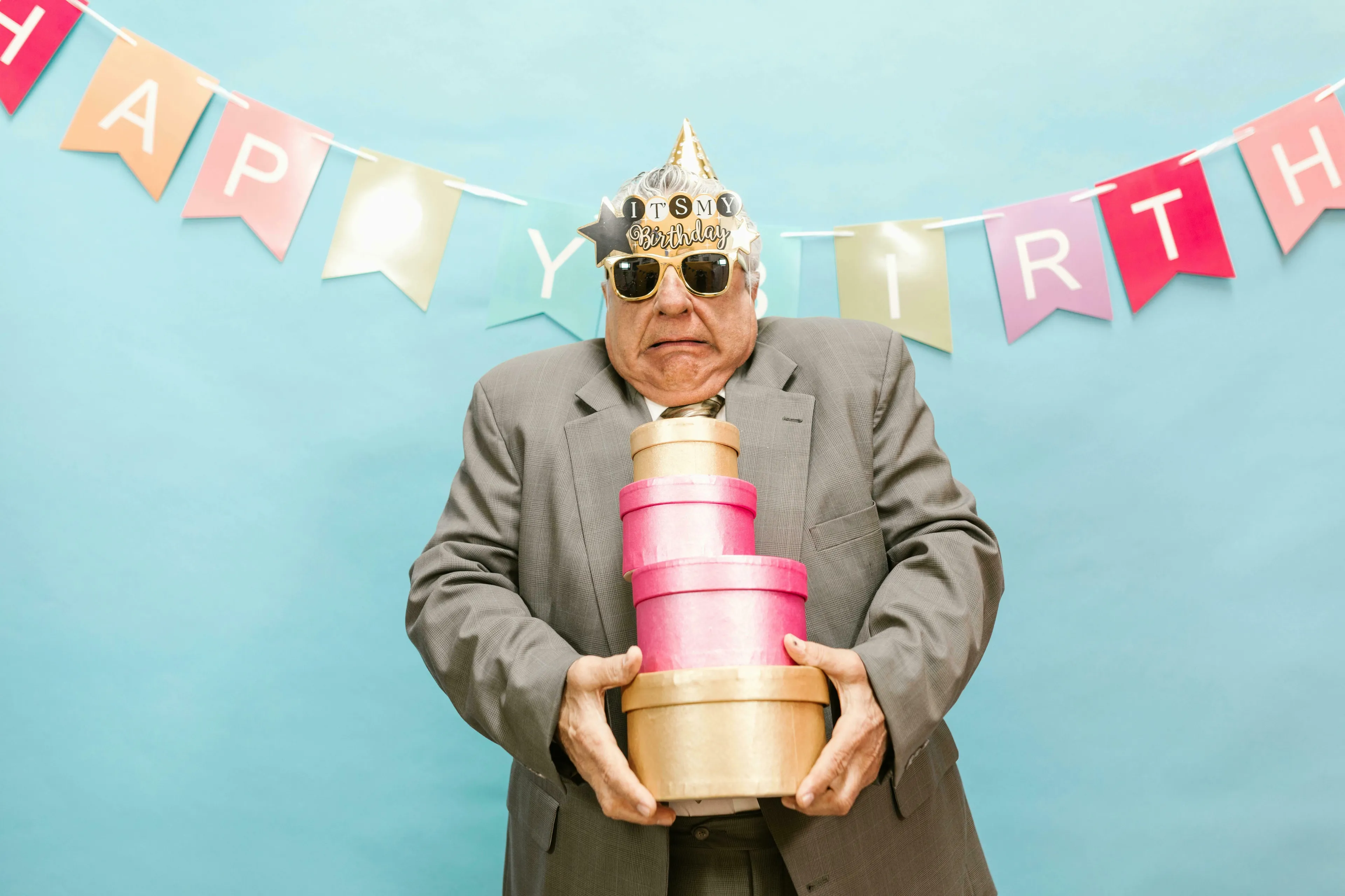 A man in a party hat and sunglasses holds gifts, under a Happy Birthday banner. Test your General Knowledge Quiz for High IQs.