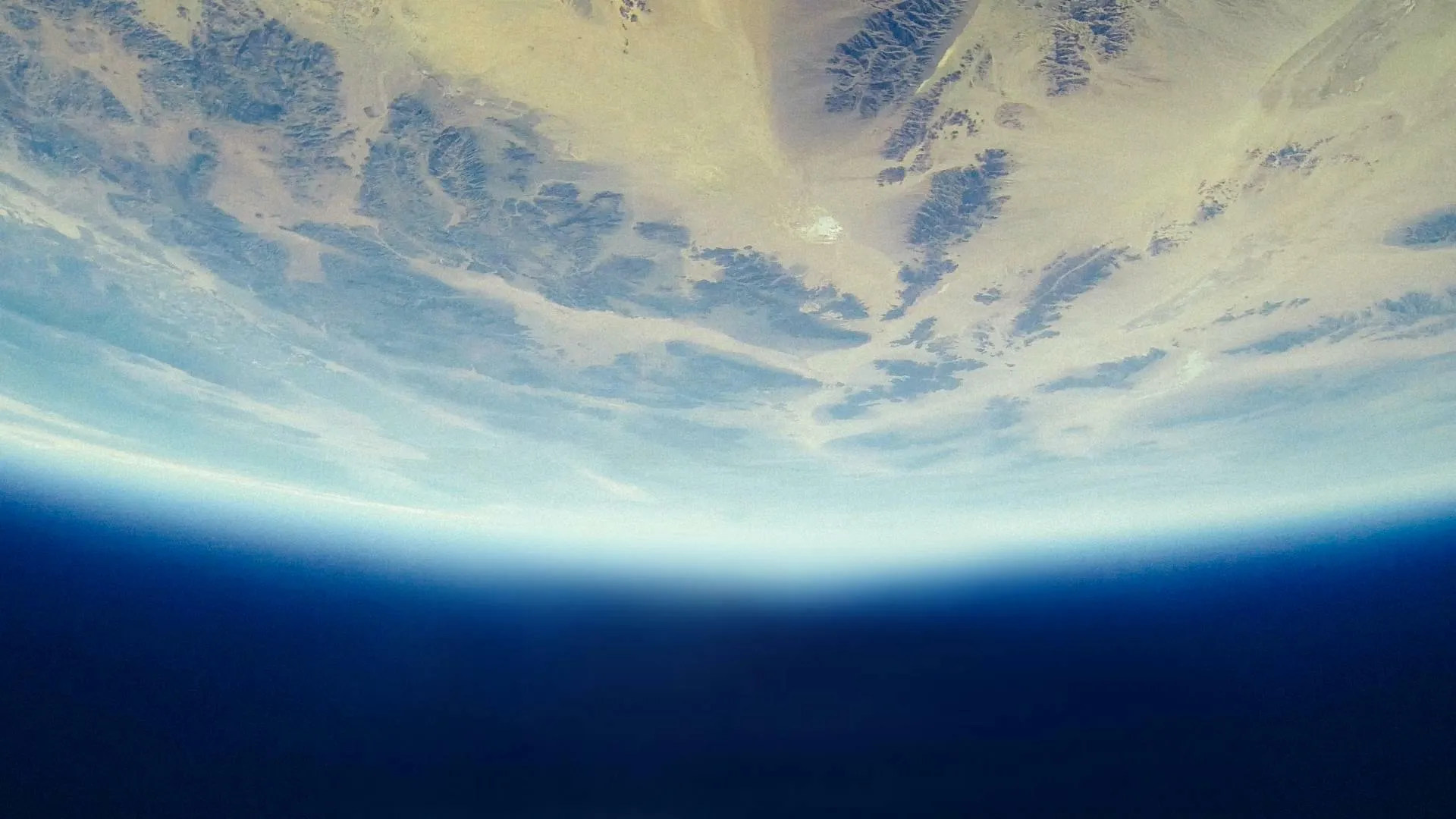 View of Earth from space, showing a curved horizon of blue ocean meeting a tan, mountainous landmass, ideal for a difficult general knowledge quiz for High IQs.