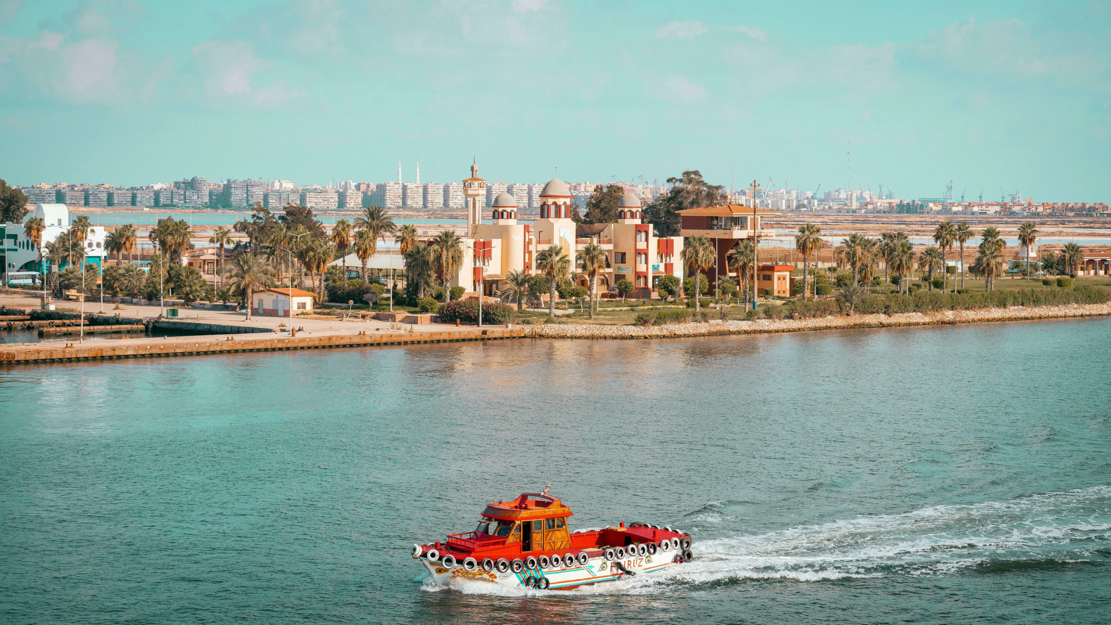 A red boat moves across the Suez Canal's turquoise waters, with buildings and palm trees lining the shore. General Knowledge Quiz.