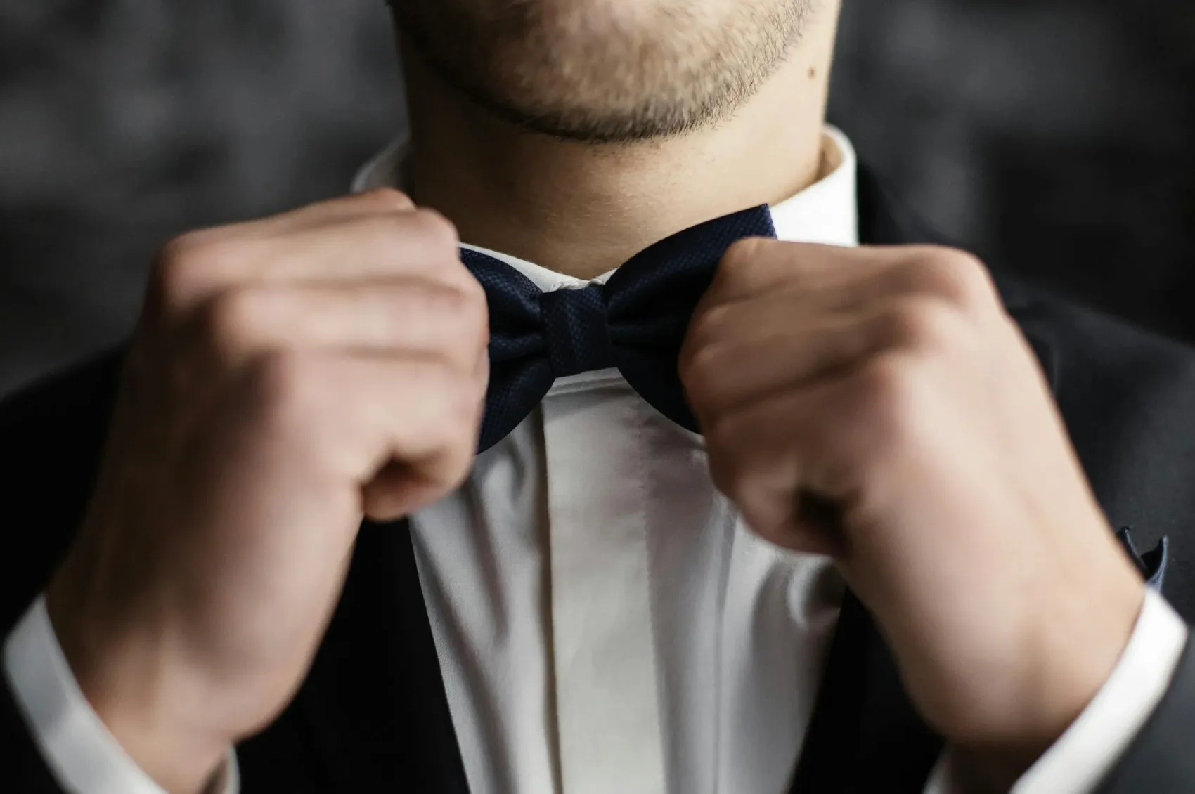 A man adjusts a navy blue bow tie, worn with a white shirt and black suit jacket. Popular Y2K accessory.