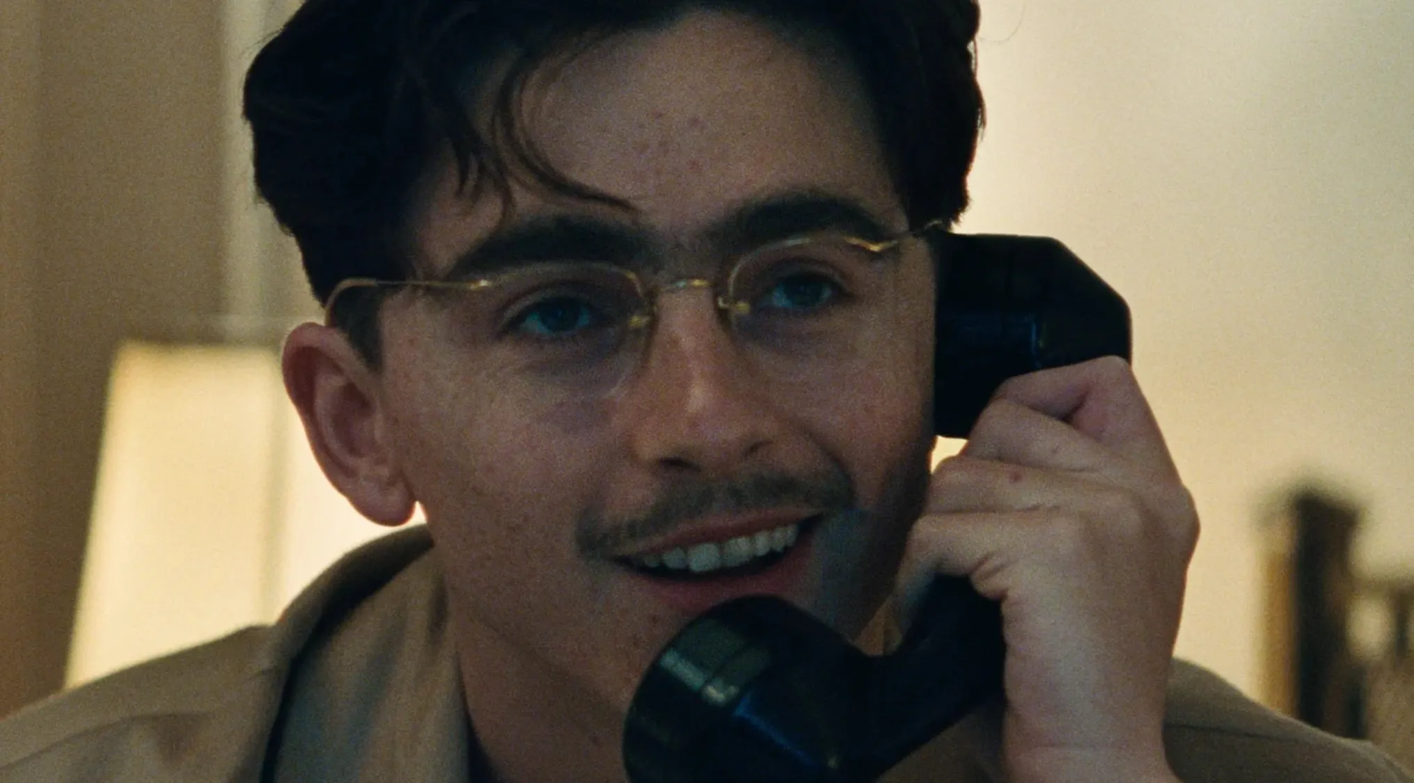 A smiling man with a mustache and glasses holding a phone, part of a quiz on how well you spell celebrity names.