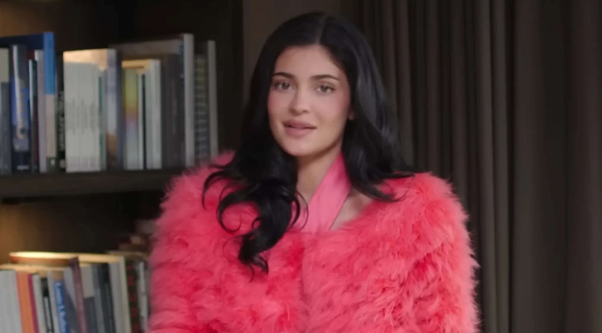 Kylie Jenner in a bright pink feather coat, against a bookshelf. Can you spell celebrity names correctly?