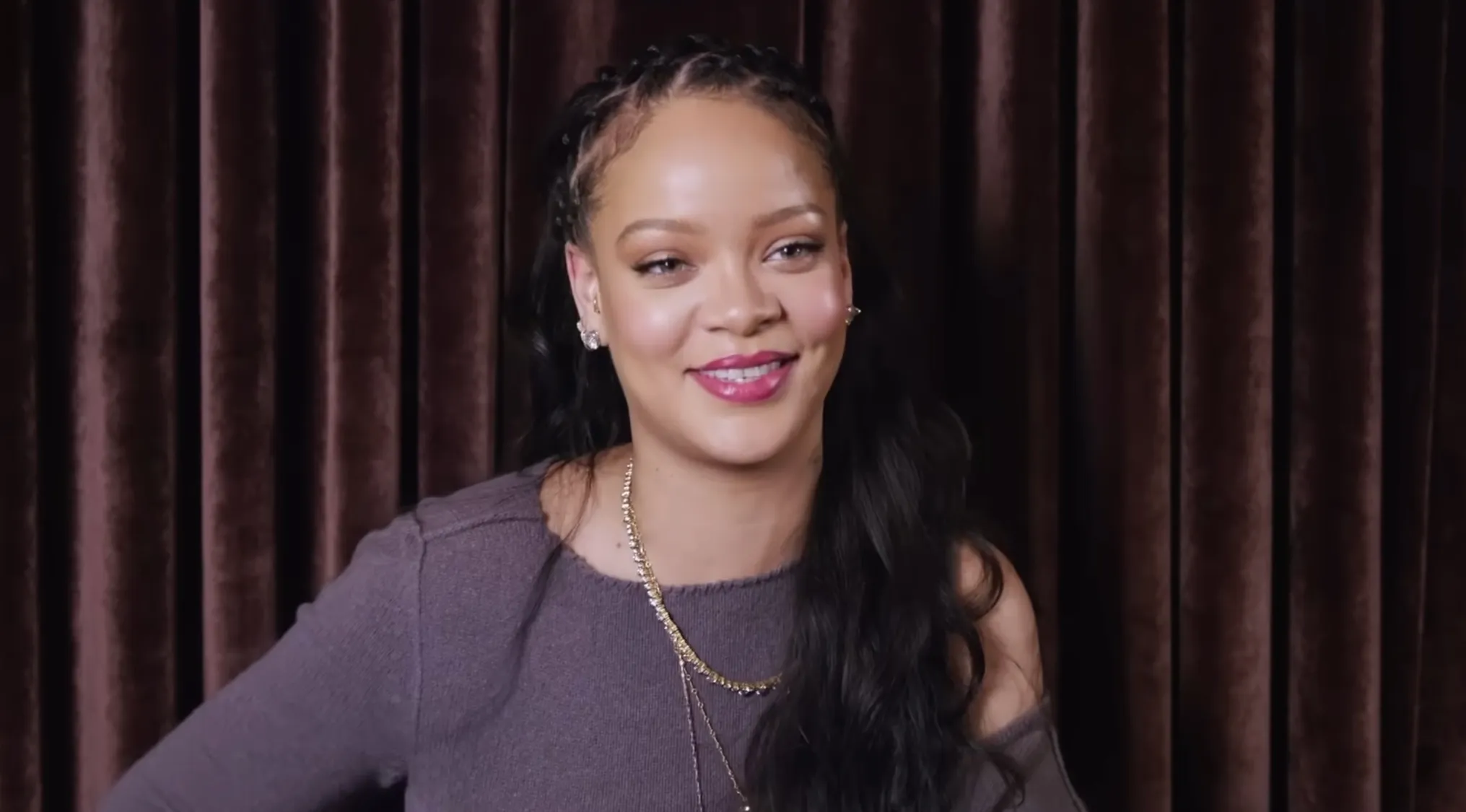 Rihanna, with braids and long dark hair, smiling against a dark brown curtain. Spell celebrity names correctly.