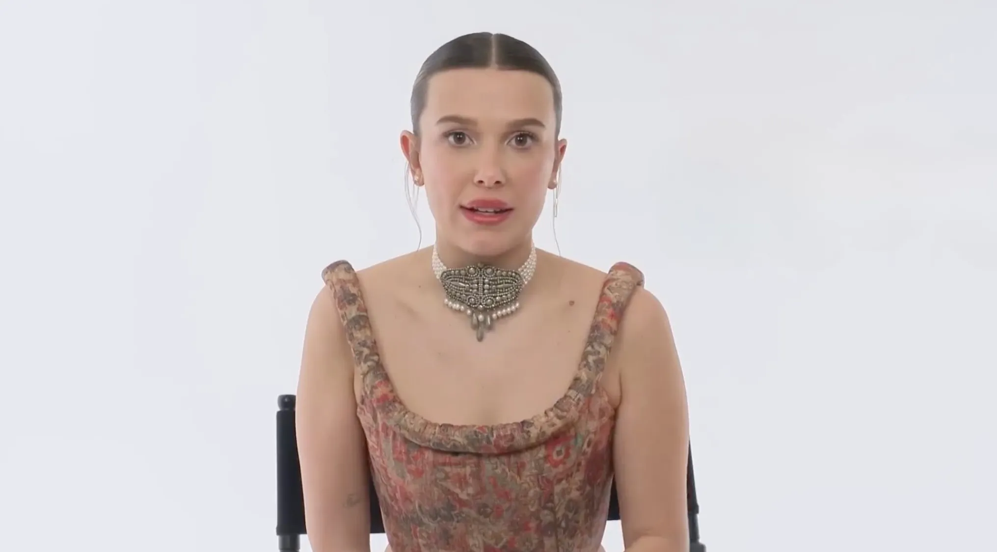 Millie Bobby Brown, spelling celebrity names. She wears a pearl choker and patterned top against a white background.