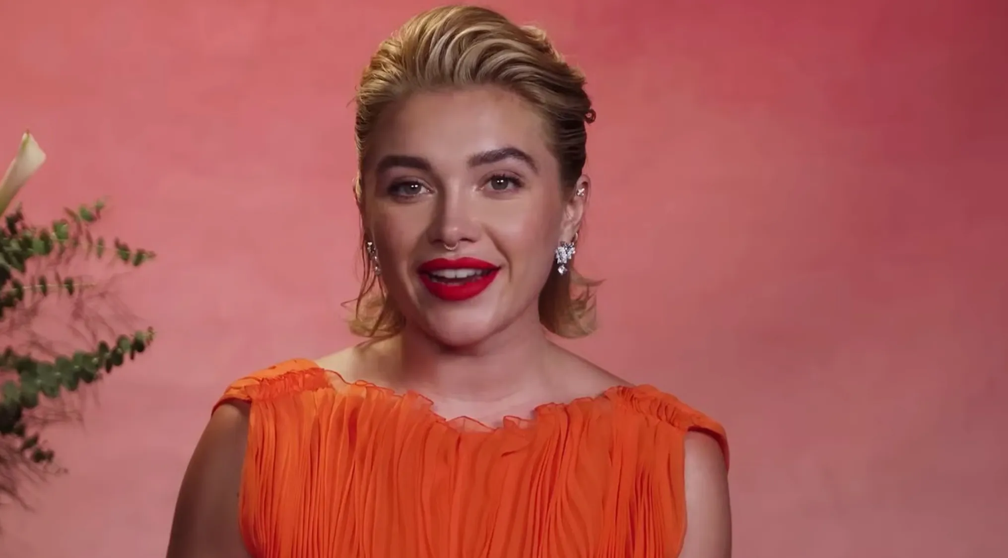 Florence Pugh in an orange dress with red lipstick, smiling. Test your ability to spell celebrity names!