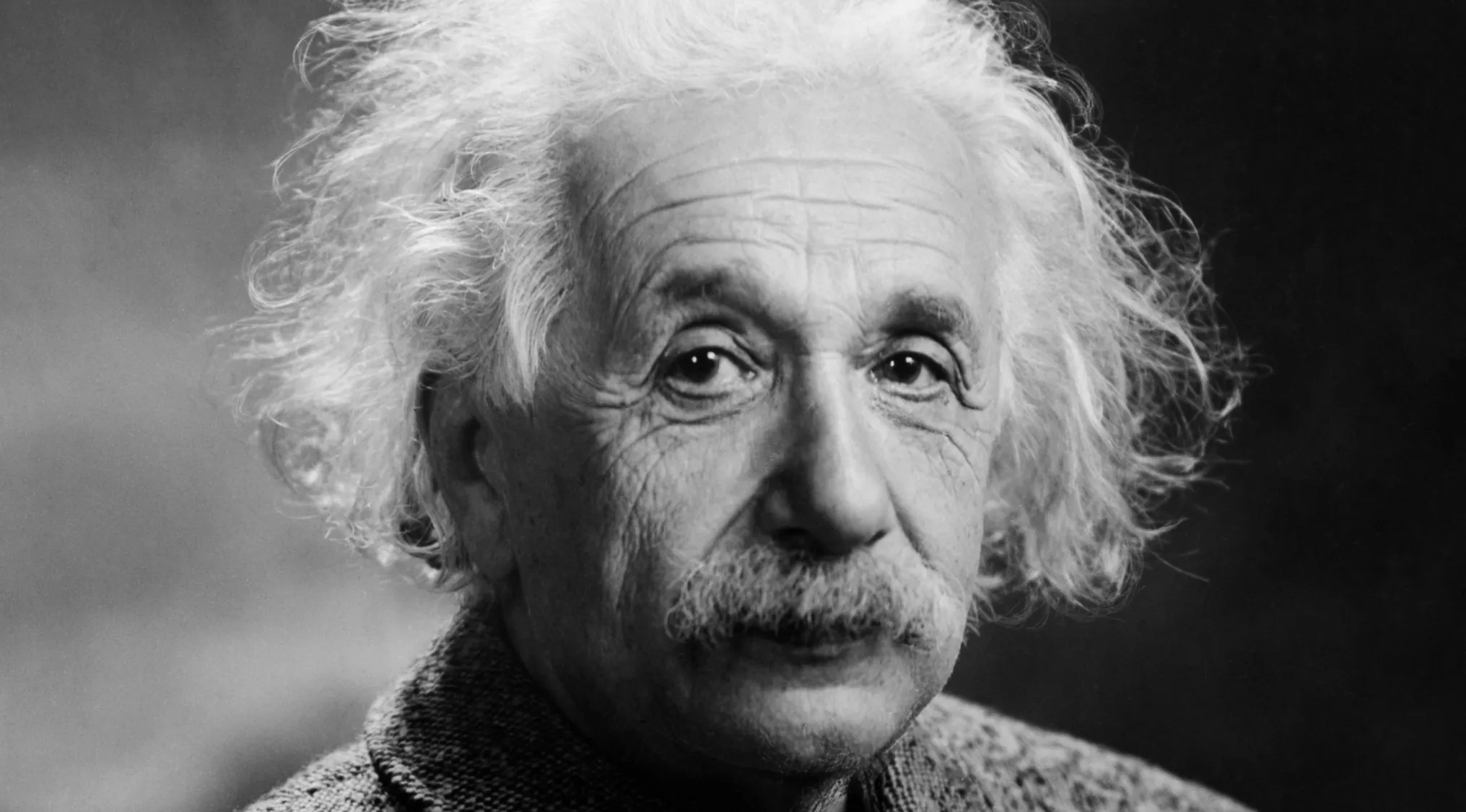 A black and white close-up of Albert Einstein's face, featuring his wild white hair and iconic mustache. Real or fake trivia.
