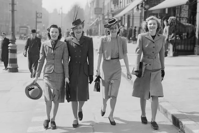 Four women in 1940s fashion, including structured suits and hats, walk confidently down a city street, smiling.