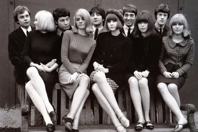 A group of young men and women in 1960s fashion sitting on a bench, looking at the camera.