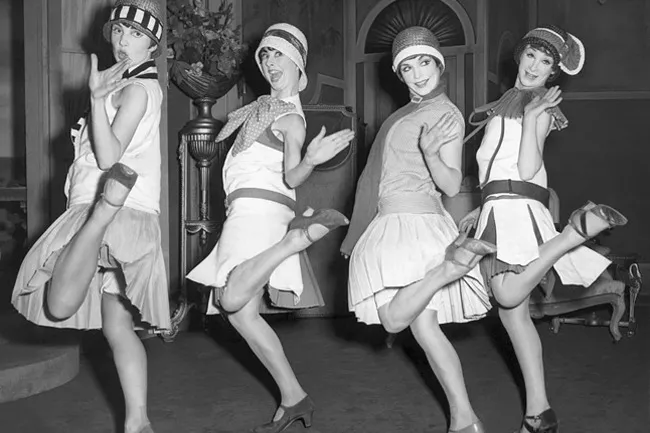 Four women in 1920s fashion, including cloche hats and short dresses, striking playful poses with their legs kicked up.