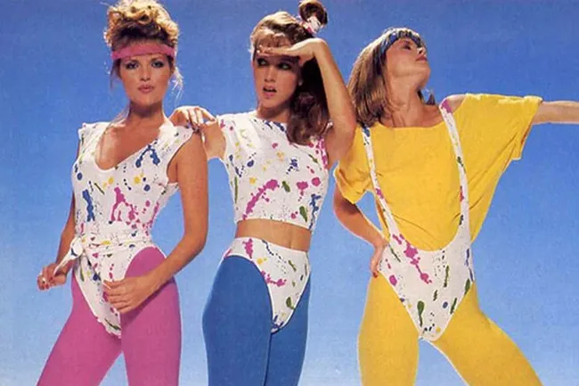 Three women in vibrant 1980s aerobics wear, featuring splash-paint designs and bright leggings, against a blue sky.