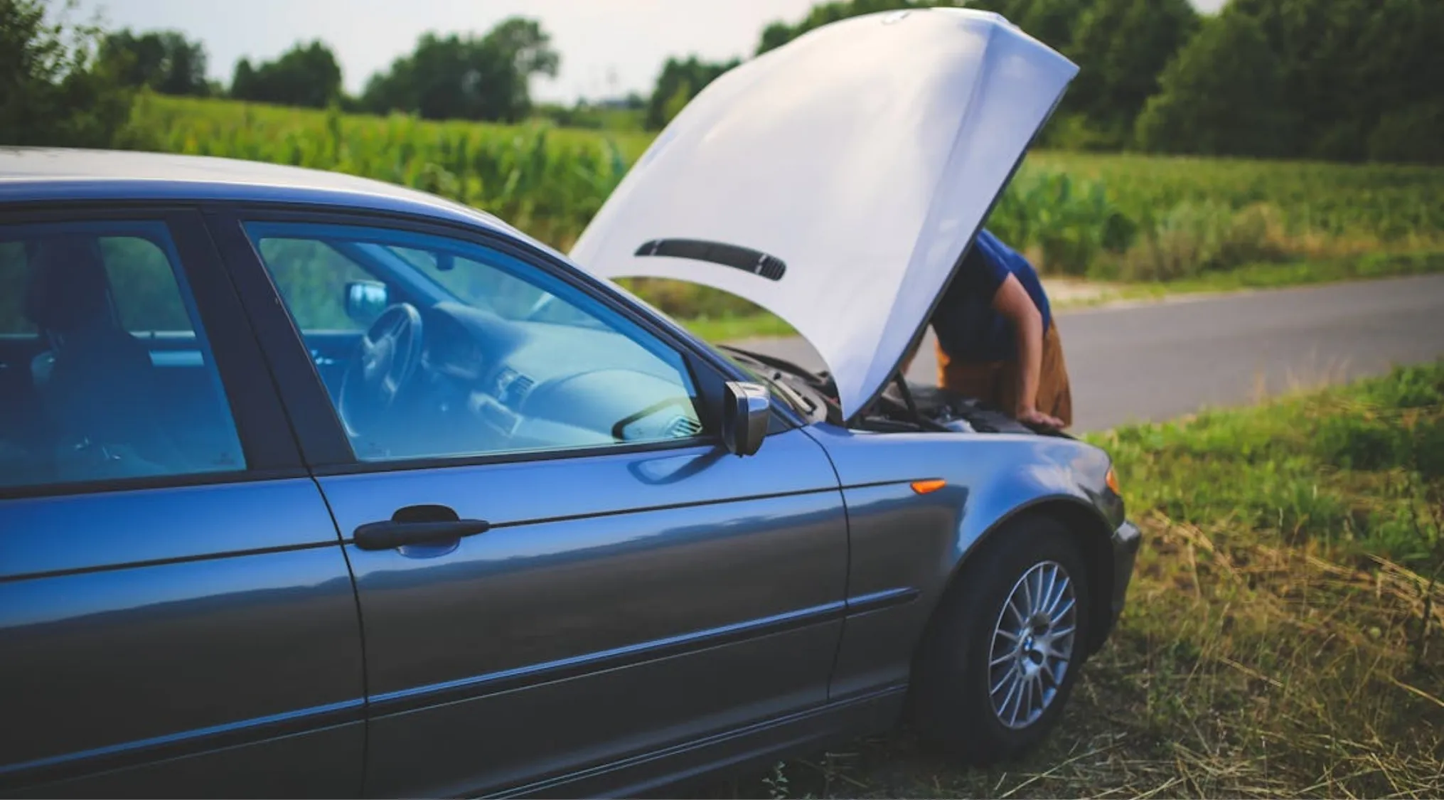 A person leaning into the open hood of a blue car on the roadside, practicing Home Economics.