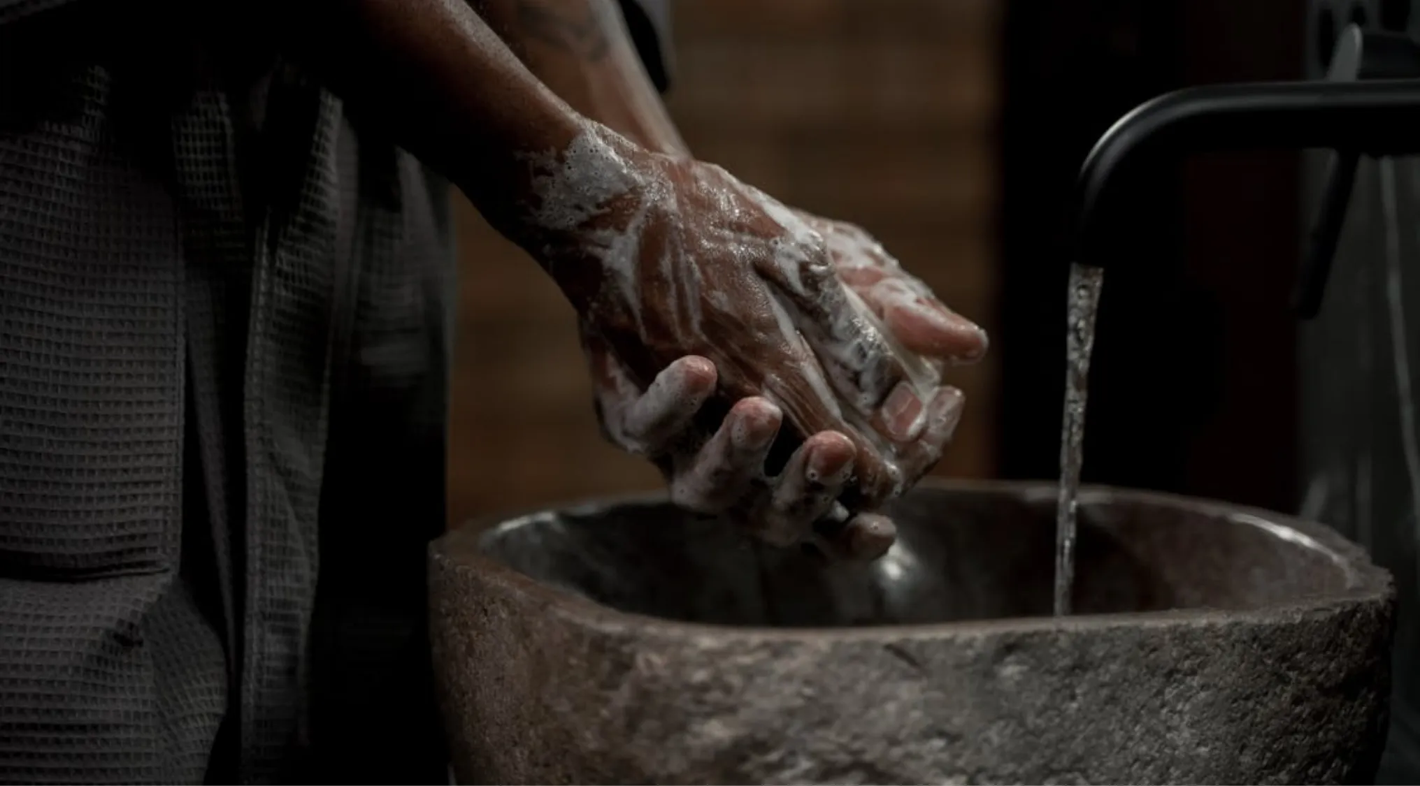 Hands covered in soap lather, being washed in a stone sink with water flowing from a black faucet. Essential for cooking hygiene.