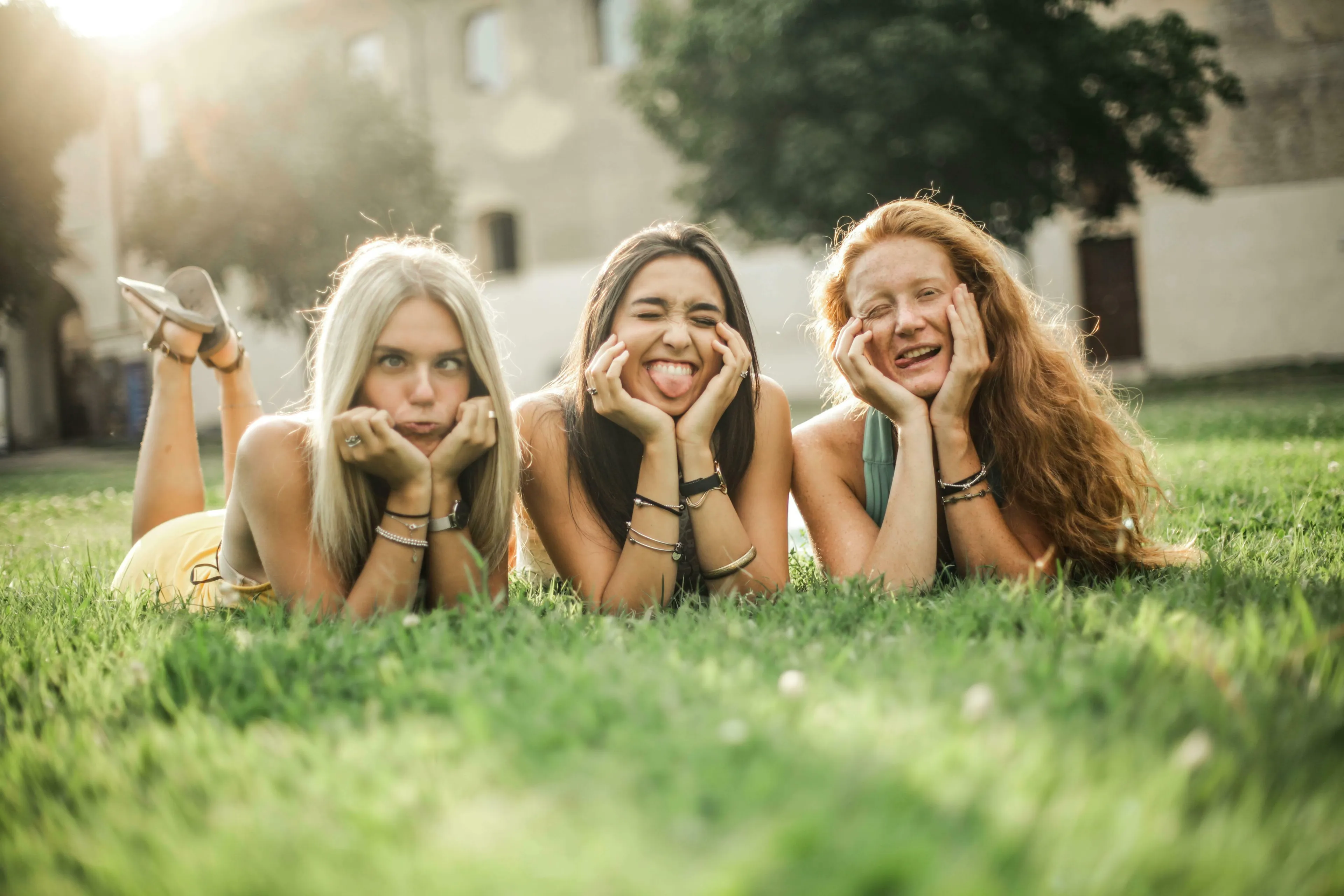 Three unique young women making funny faces while lying on green grass, enjoying the sun.