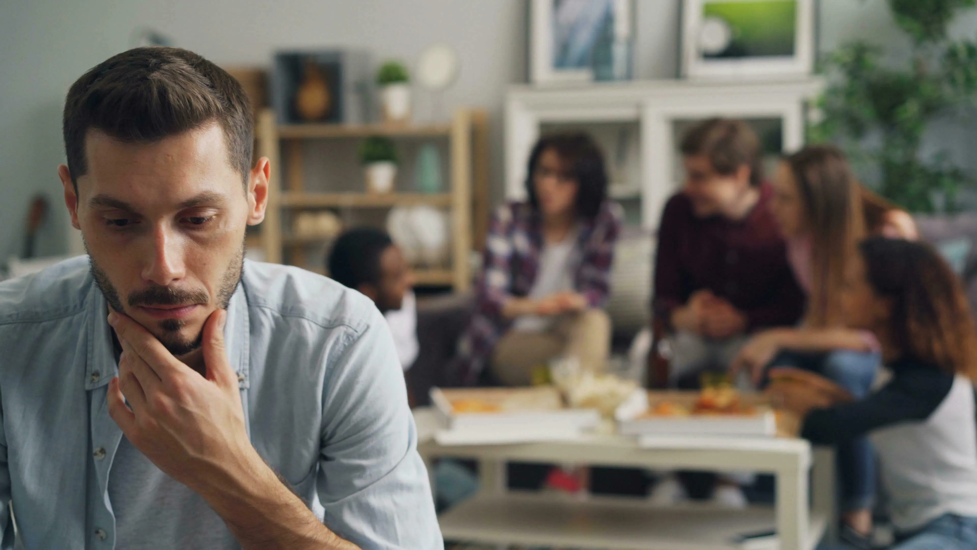 A man with a beard looks down thoughtfully, his hand on his chin. In the blurry background, a group of people socializes. Unique and mistake.