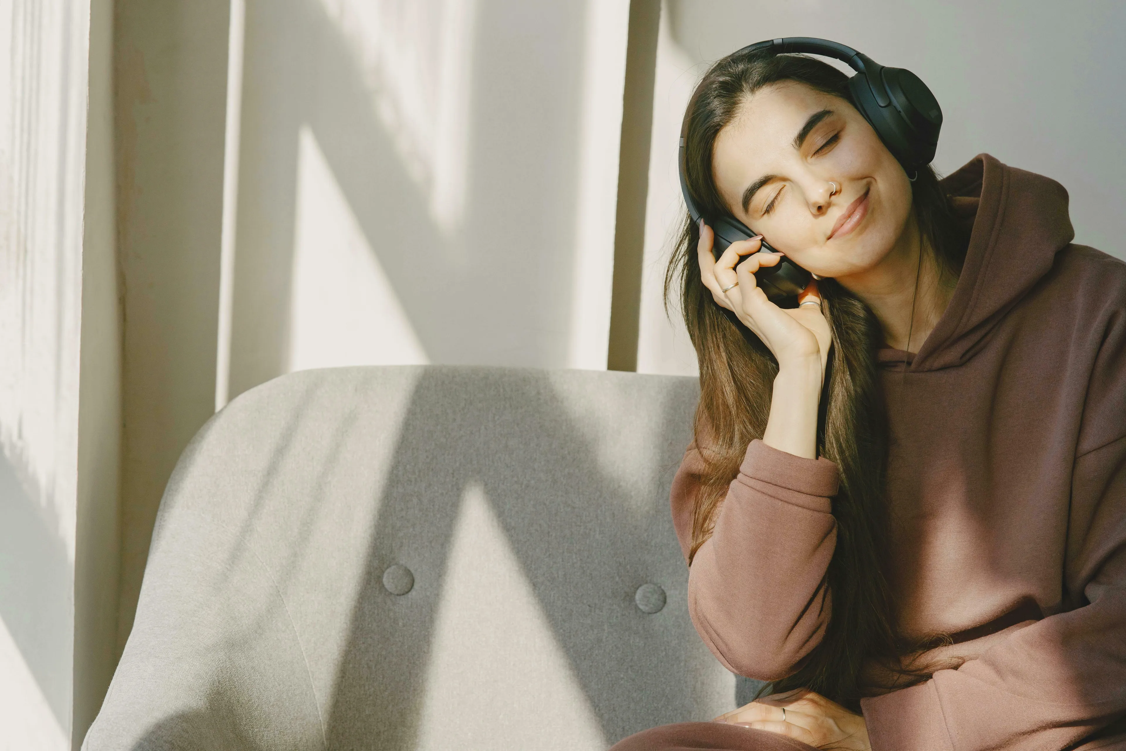 A woman with long hair, wearing headphones and a brown hoodie, smiling with eyes closed, enjoying music. How unique are you? Pick a music genre.