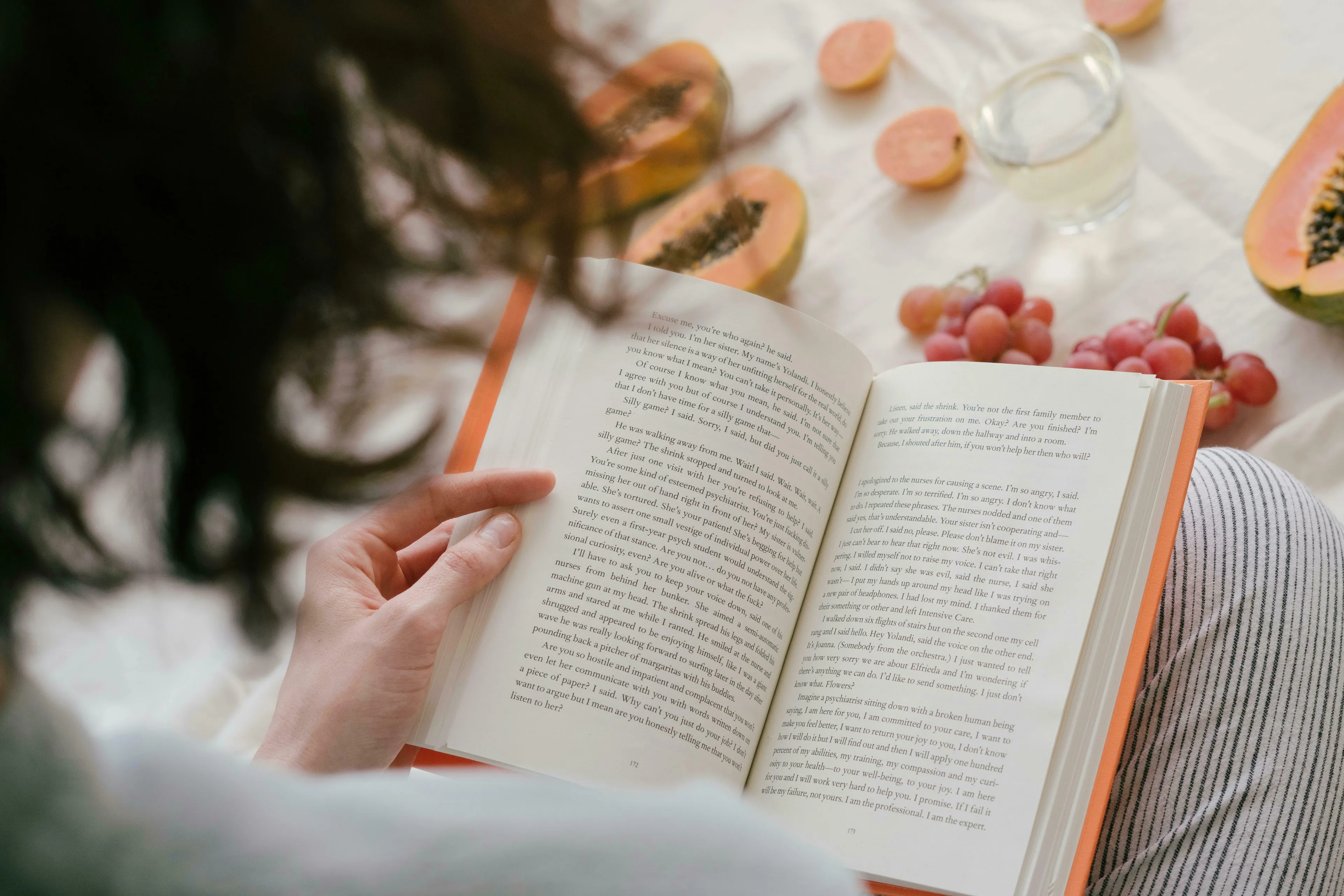 A person reads a book while relaxing, with papayas, guavas, and grapes on a white surface. The reader is unique.