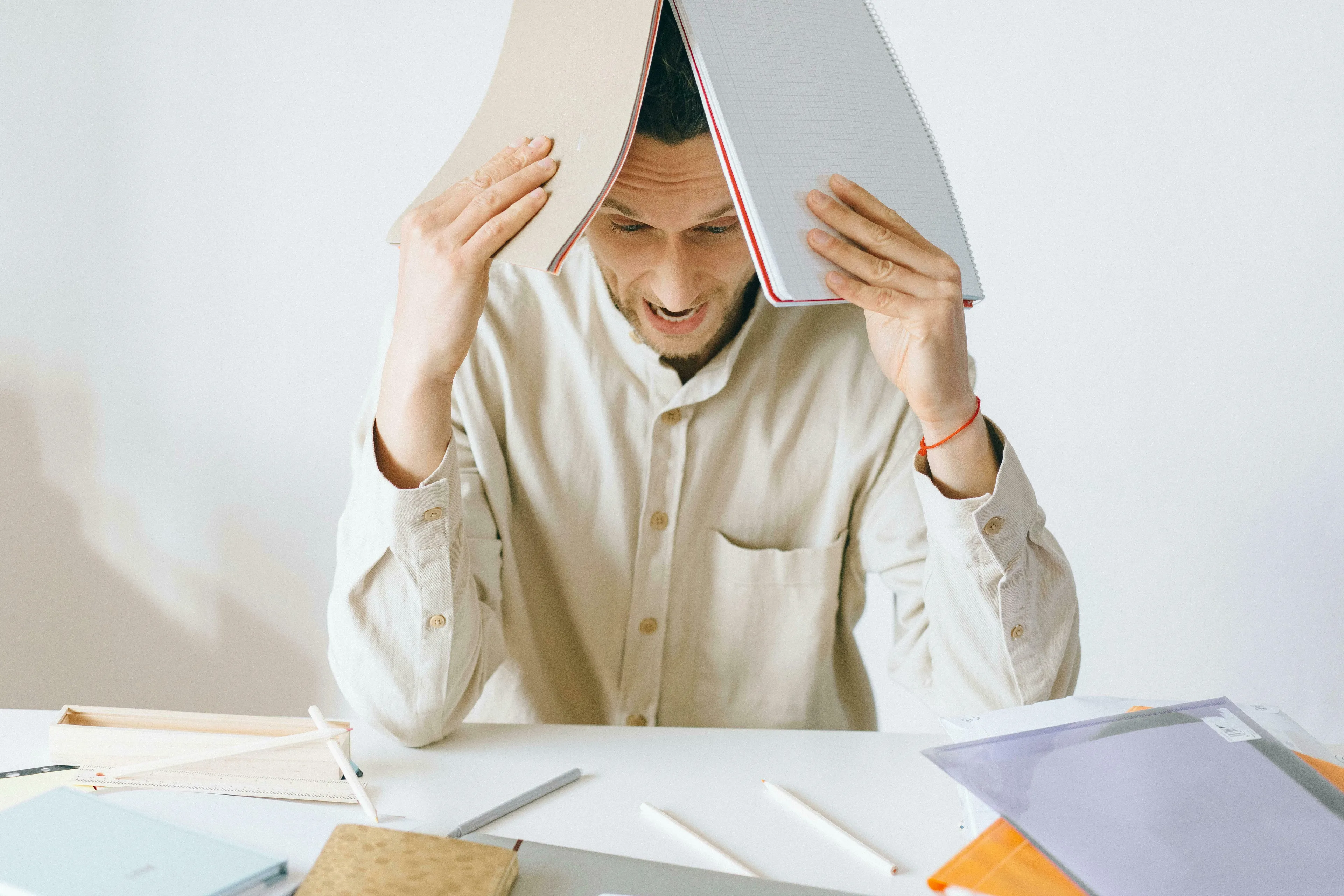 A man, overwhelmed, covering his head with notebooks at a desk. Highlights the difficulty of making a big decision and how unique you are.