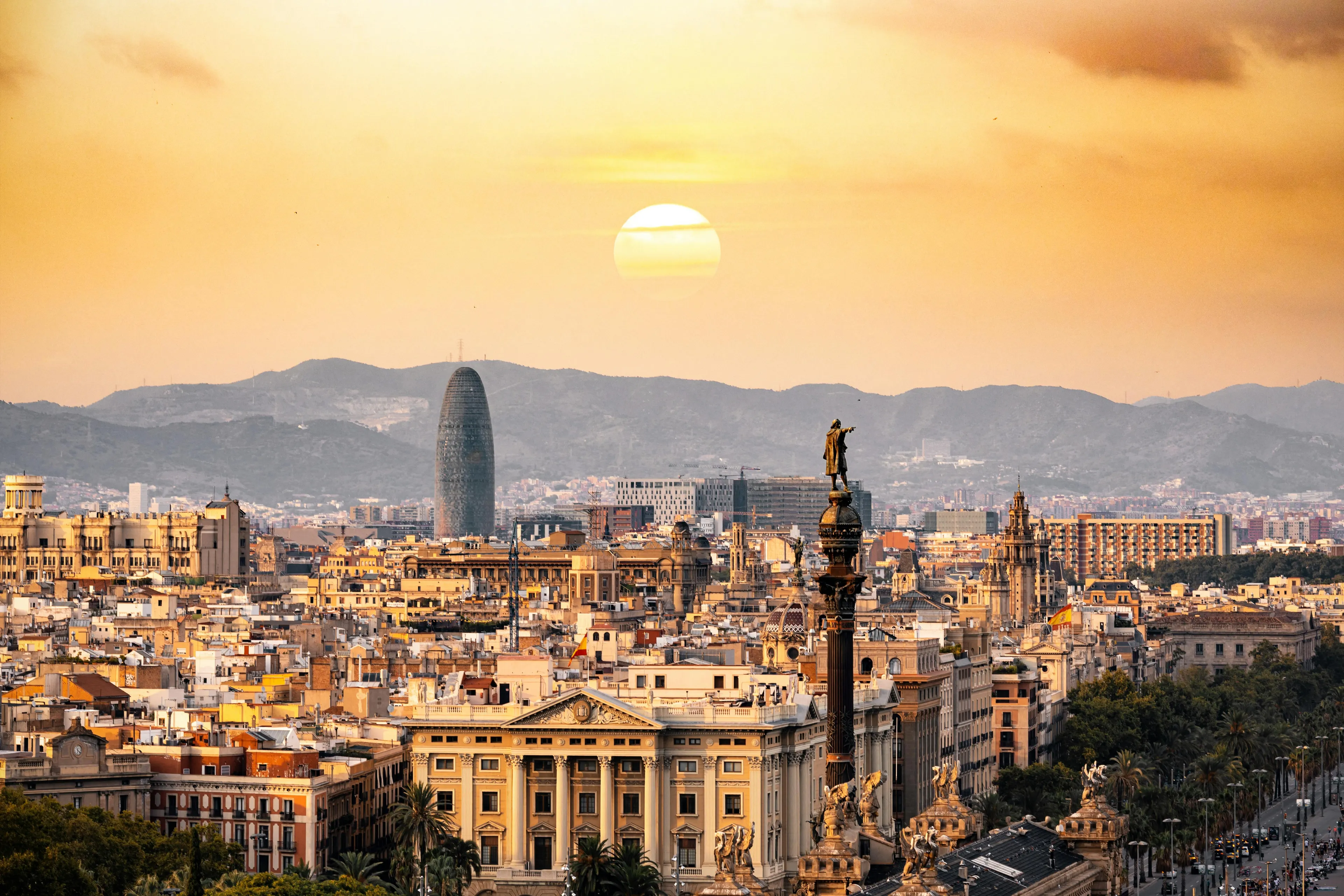 Aerial view of the Barcelona cityscape at sunset, with historical buildings, a distinctive skyscraper, and mountains in the distance.