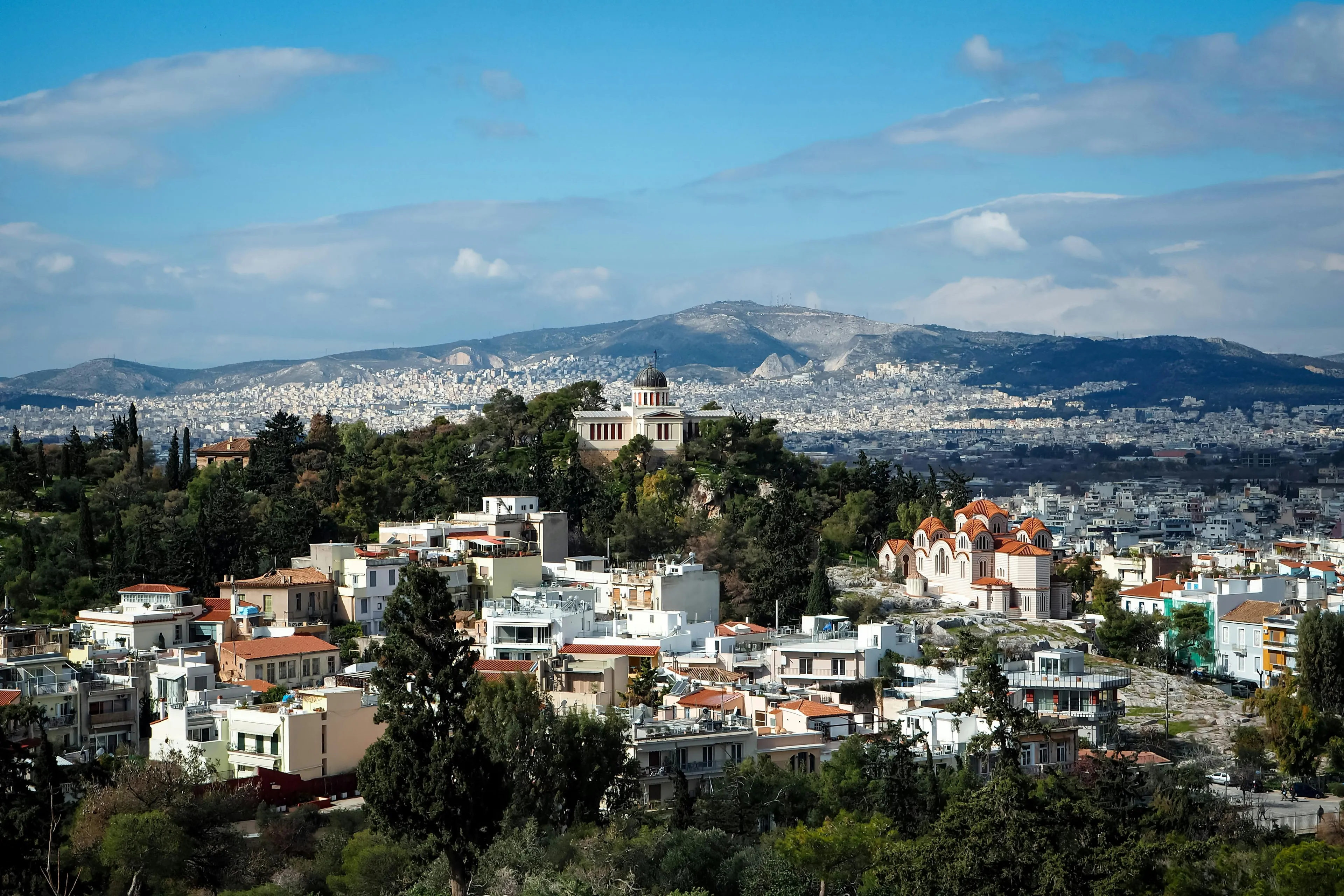 An elevated view of Athens, Greece, showcasing white and terracotta buildings nestled among green trees, with distant mountains. Long weekend.