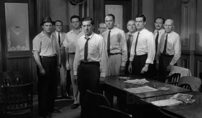 The 12 Angry Men jury stands in a black and white film still, dressed in shirts and ties, gathered in a deliberation room.