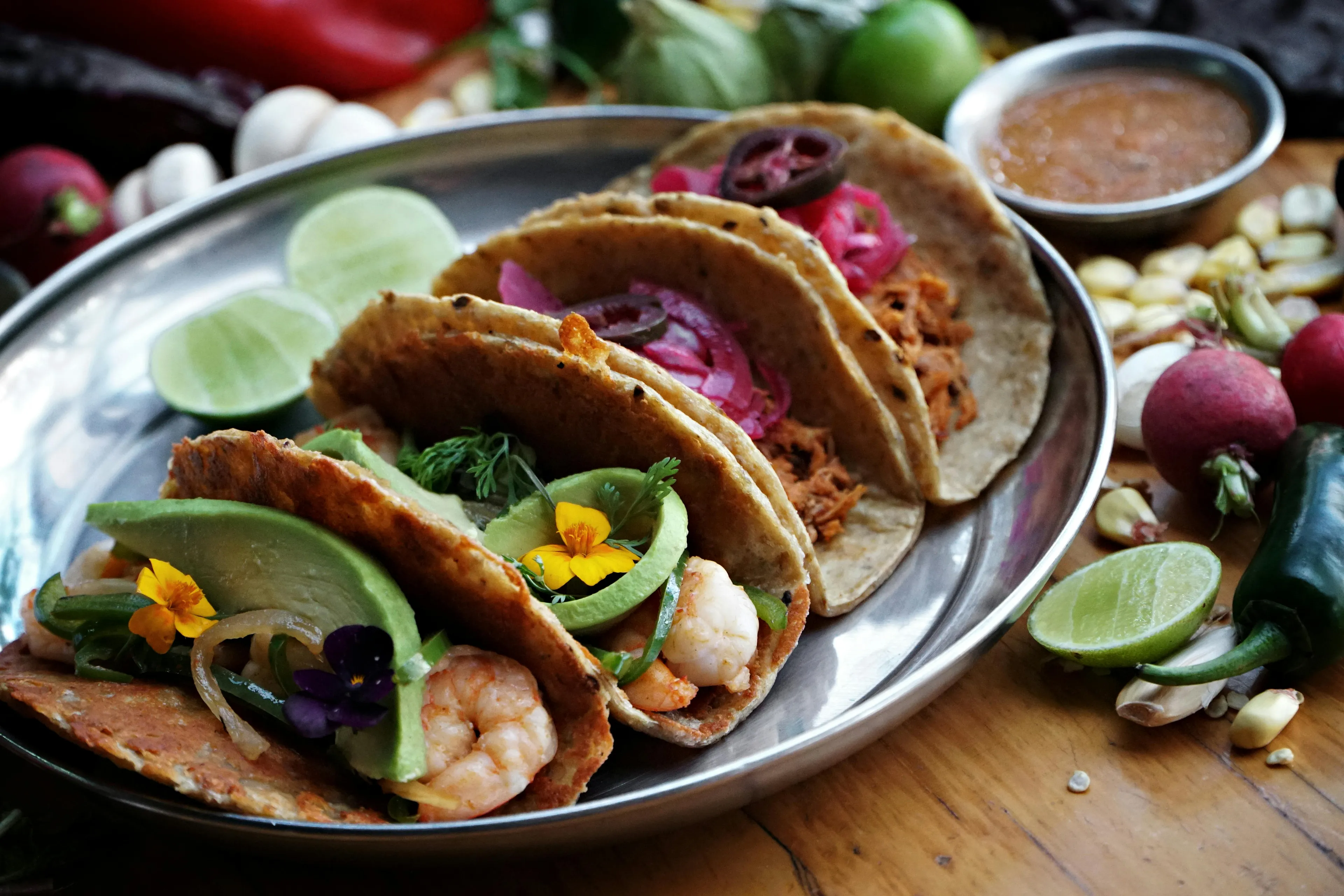 A vibrant plate of various tacos, including shrimp and pulled pork, adorned with fresh avocado and edible flowers. A perfect meal.