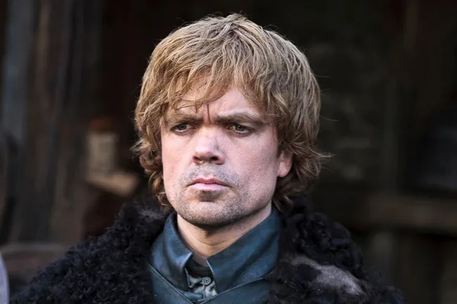 A close-up of Tyrion Lannister with short blonde hair and a beard, wearing a black fur coat.