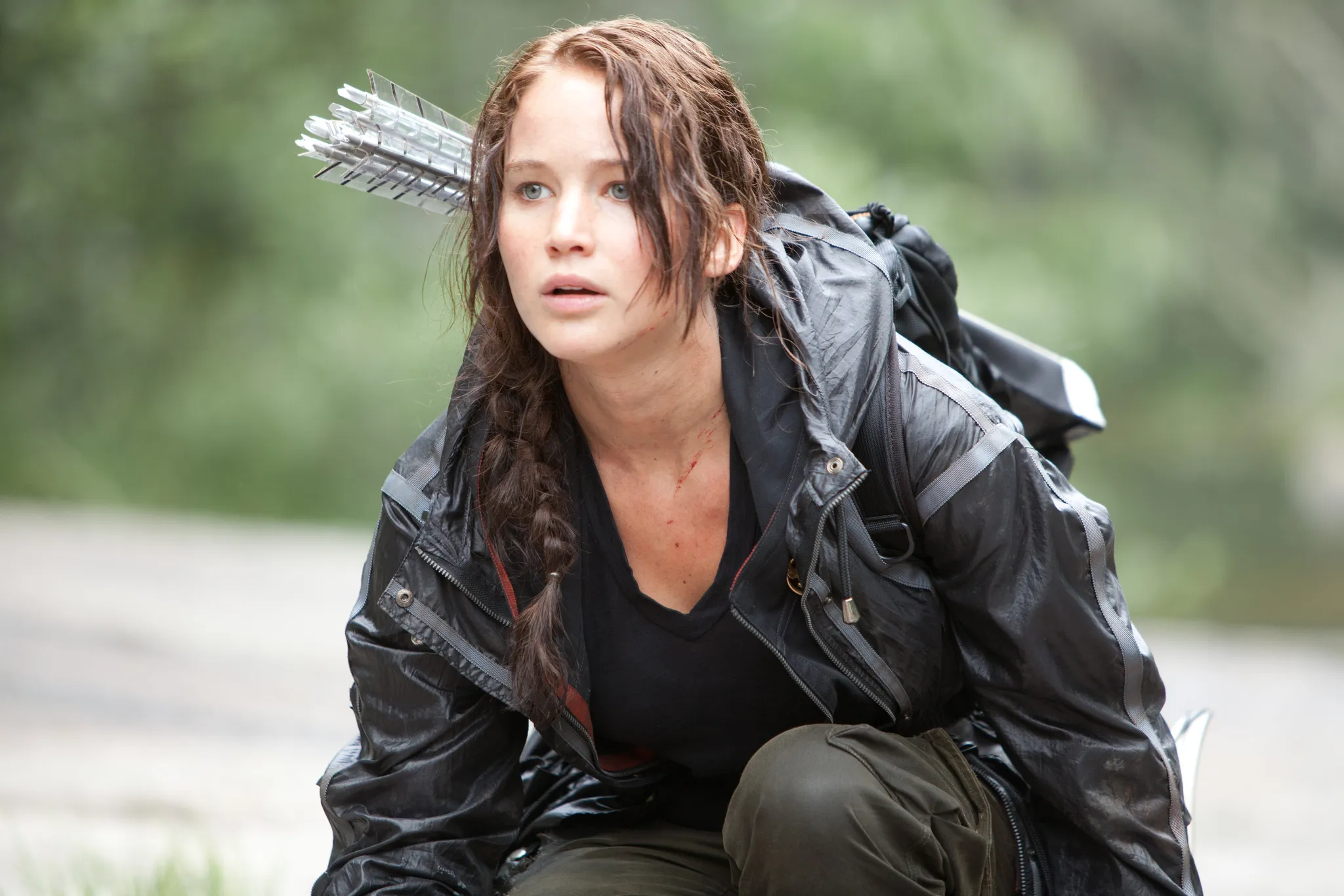 Katniss Everdeen, with her iconic braid and quiver of arrows, looking intently forward while crouching outdoors.