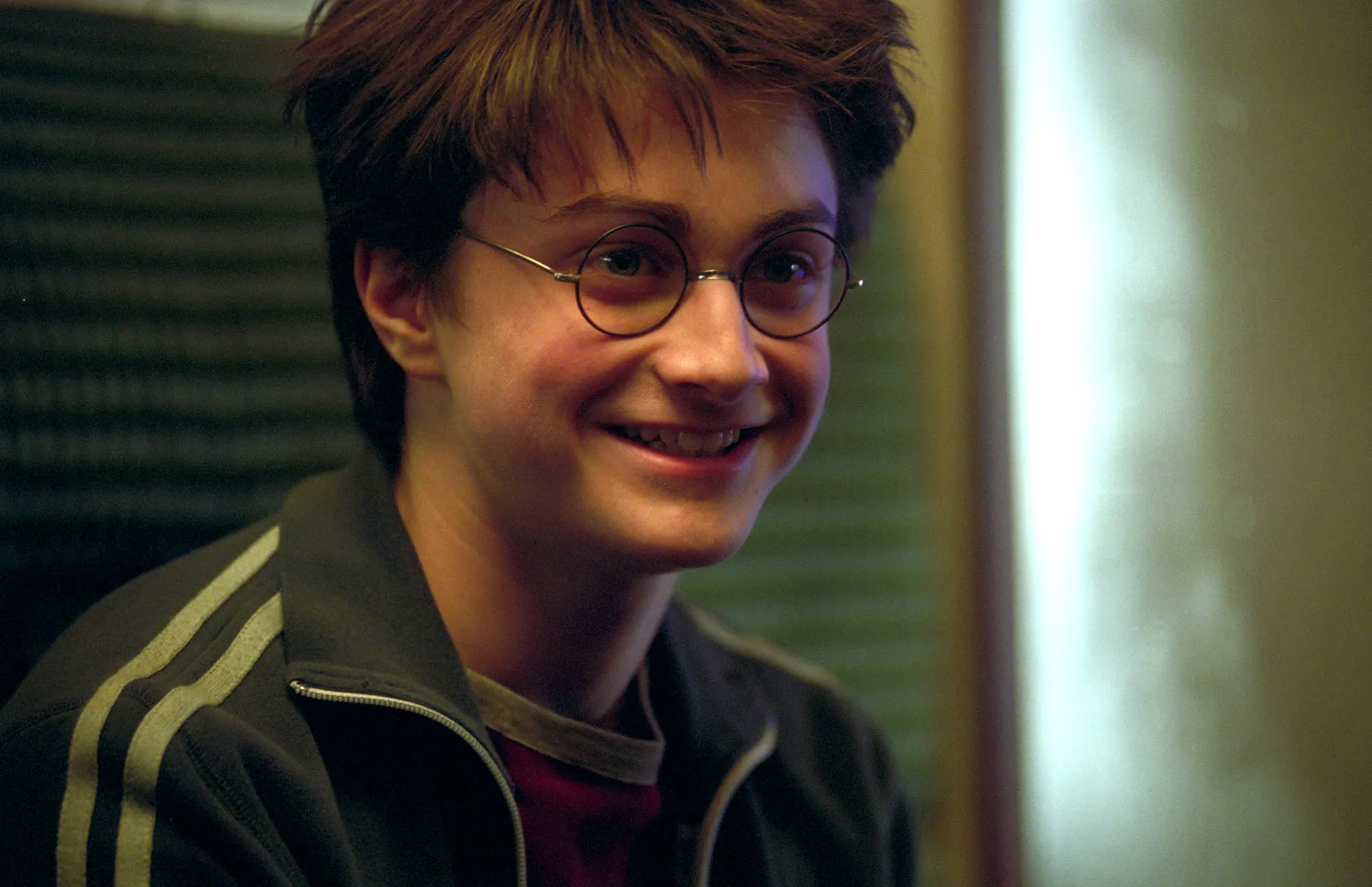 Young Harry Potter, with messy brown hair and round glasses, smiling genuinely while wearing a striped jacket.