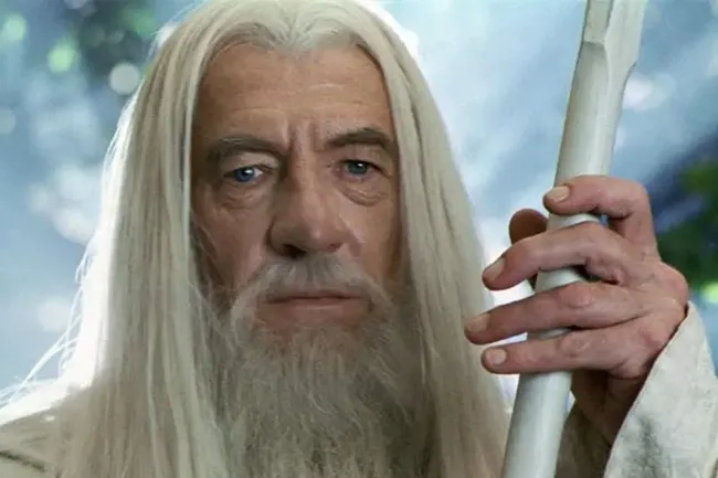Close-up of Gandalf, an older man with long white hair and beard, holding a staff, looking intently at the viewer.