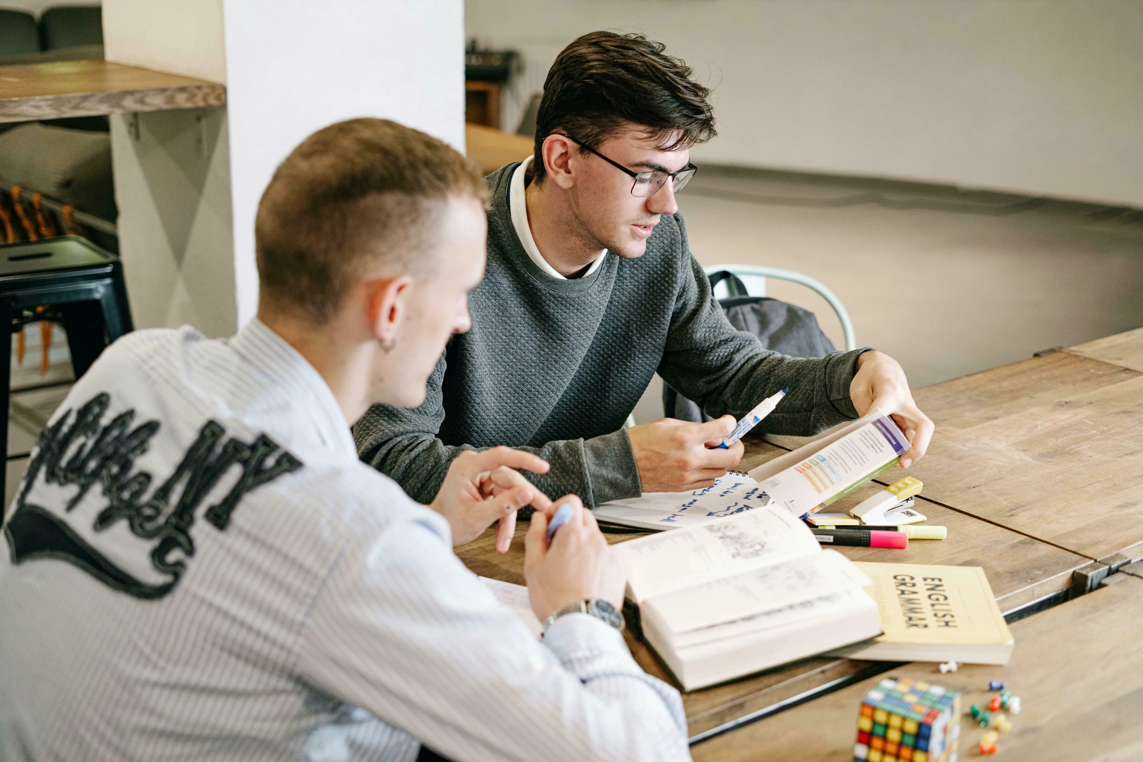 Two men studying at a wooden table, one with glasses, holding a pen. They are focused on learning which word is correctly written.