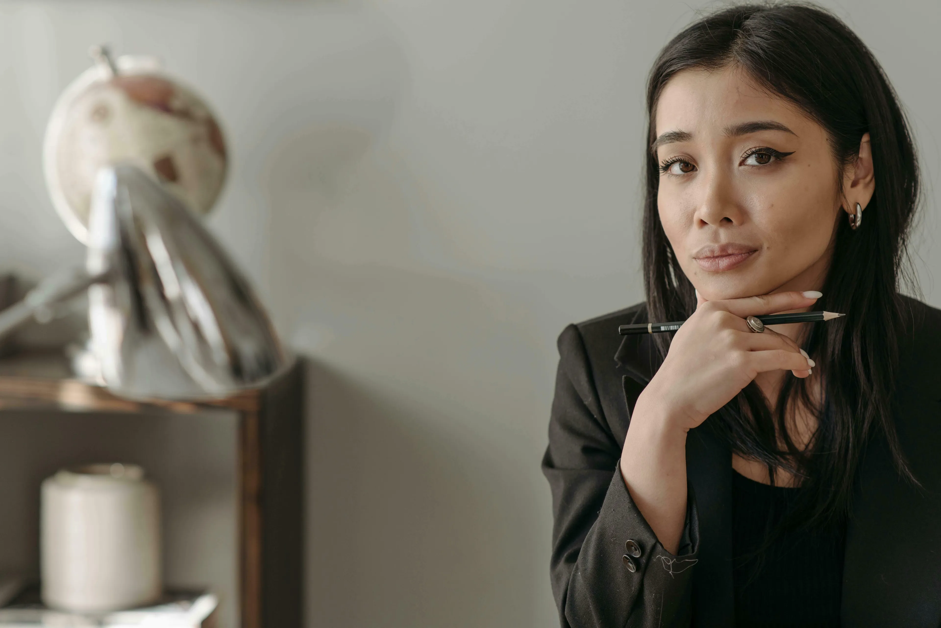 A woman with dark hair in a black blazer, holding a pencil, with a thoughtful expression. Anemology.