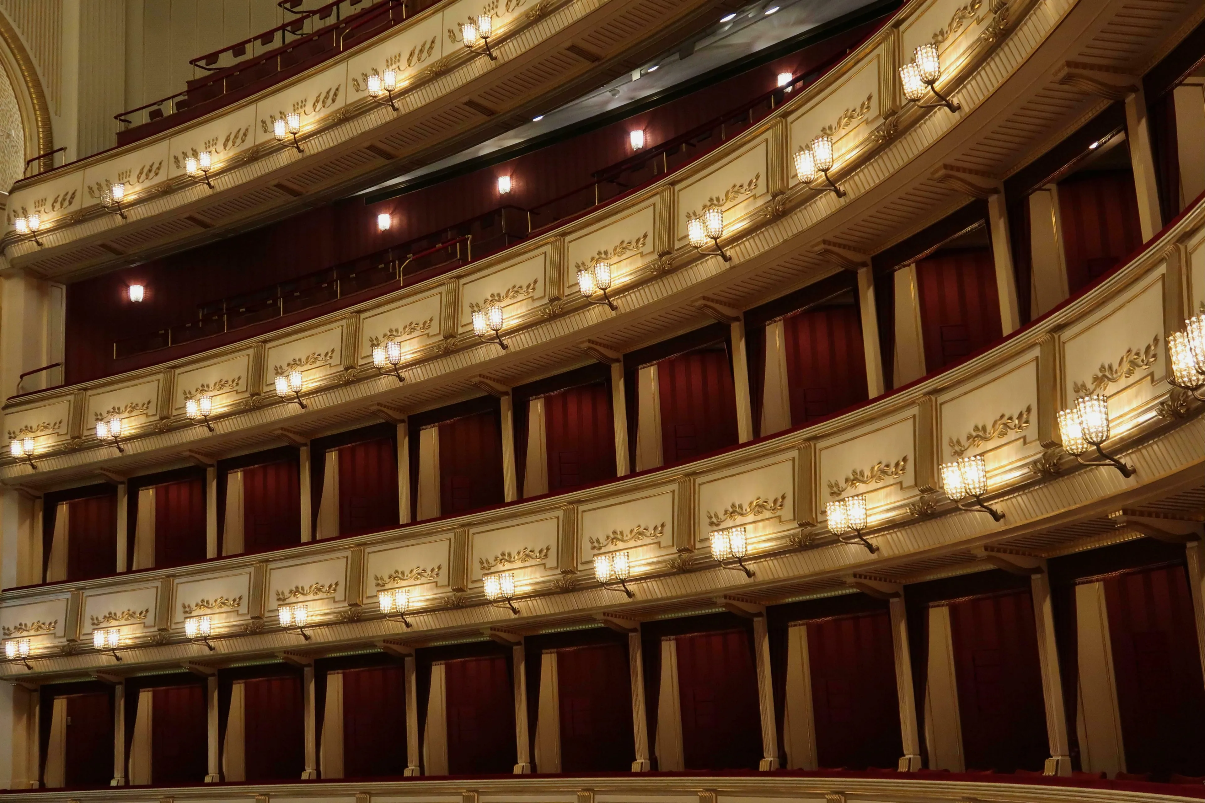 An opulent opera house interior with multiple curved tiers of private boxes, red curtains, and ornate golden wall sconces. The Marriage of Figaro.