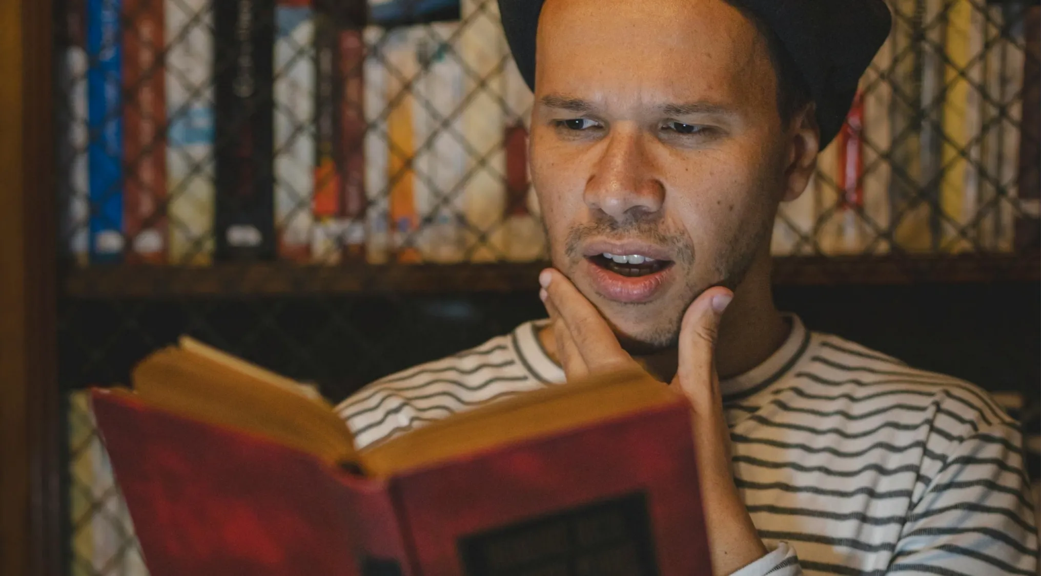 A man in a striped shirt reads a red book, looking surprised. He's thinking about General Literature Trivia.