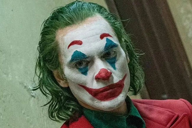 The Joker, Joaquin Phoenix, in full clown makeup with green hair, a red suit, and a serious expression.