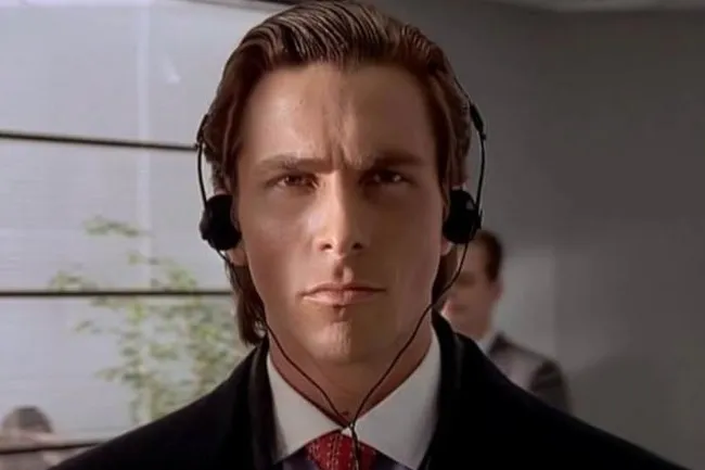 Patrick Bateman in a suit and tie, wearing headphones, looking intently at the viewer. A villain with an intense gaze.