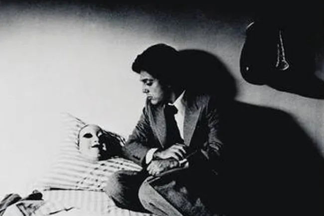 Black and white photo of Billy Joel from his Vienna song. He sits with a mask on a striped pillow, dark shadows around.