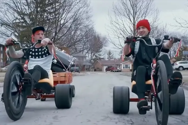 Twenty One Pilots band members on custom tricycles, one carrying drums, riding down a suburban street, feeling Stressed Out.