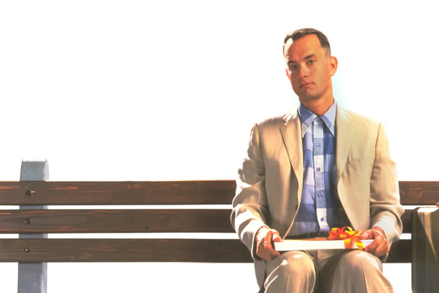 Tom Hanks as Forrest Gump sits on a bench, holding a box of chocolates. He wears a suit, from the Best Picture winning film.