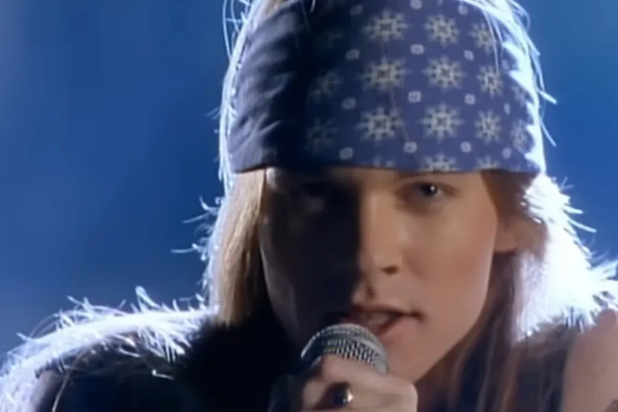 Axl Rose, with long hair and a blue bandana, singing into a microphone under bright stage lights. Lead singer.