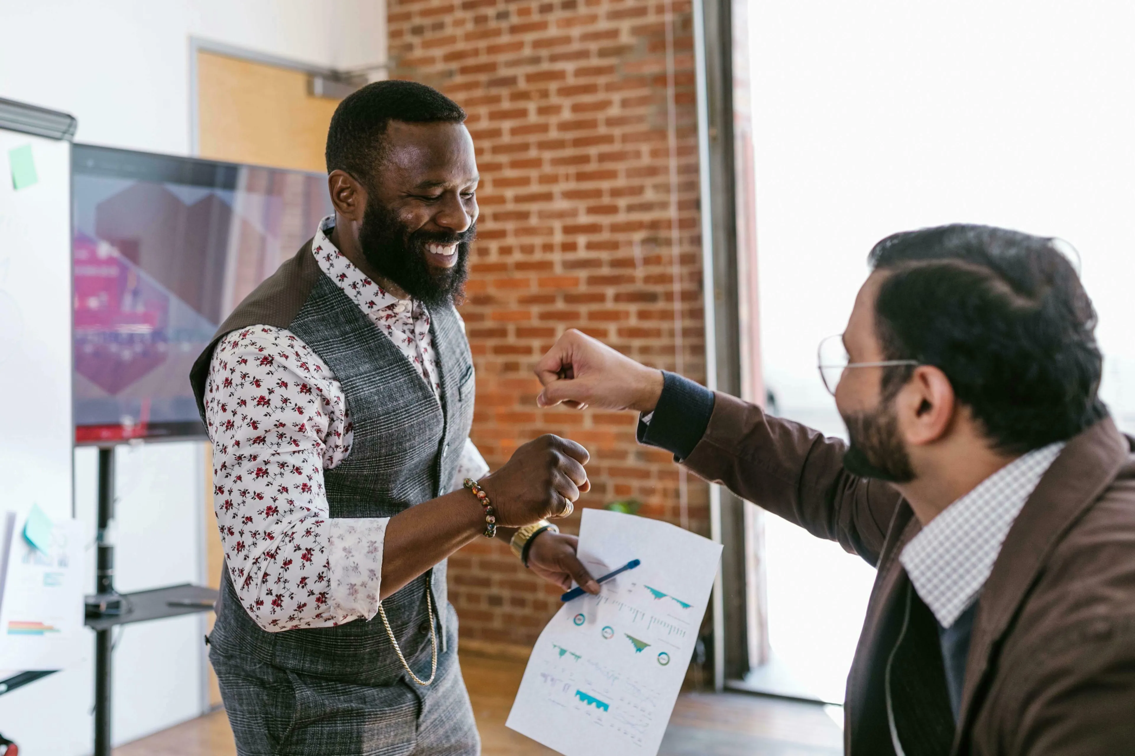 Two smiling men fist-bumping. One holds a document with charts. Reflects on procrastination type and reacting when someone finishes something before you.