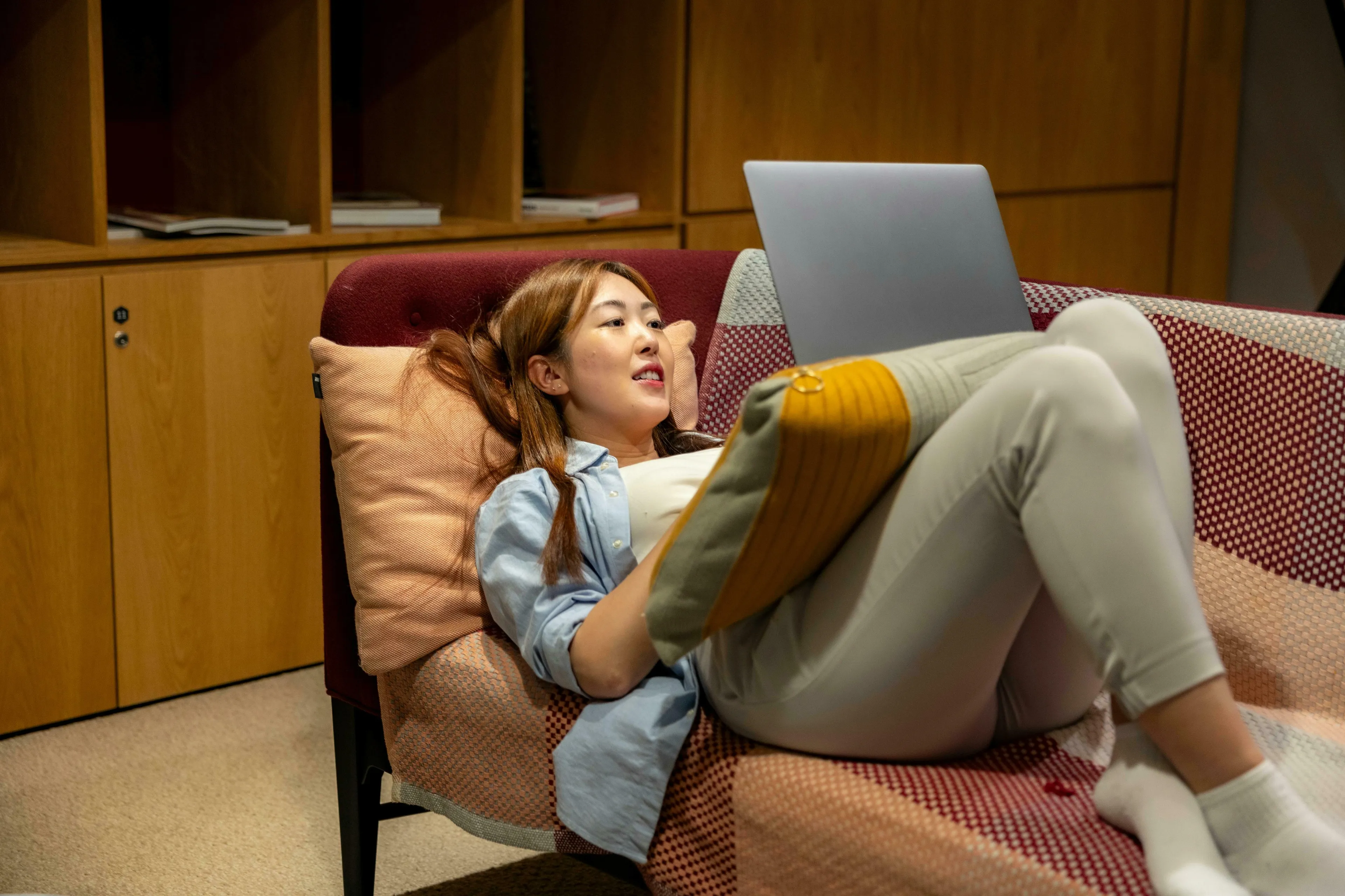 A young Asian woman lounging comfortably on a sofa, using a laptop. Discover your procrastination type and ideal work environment.