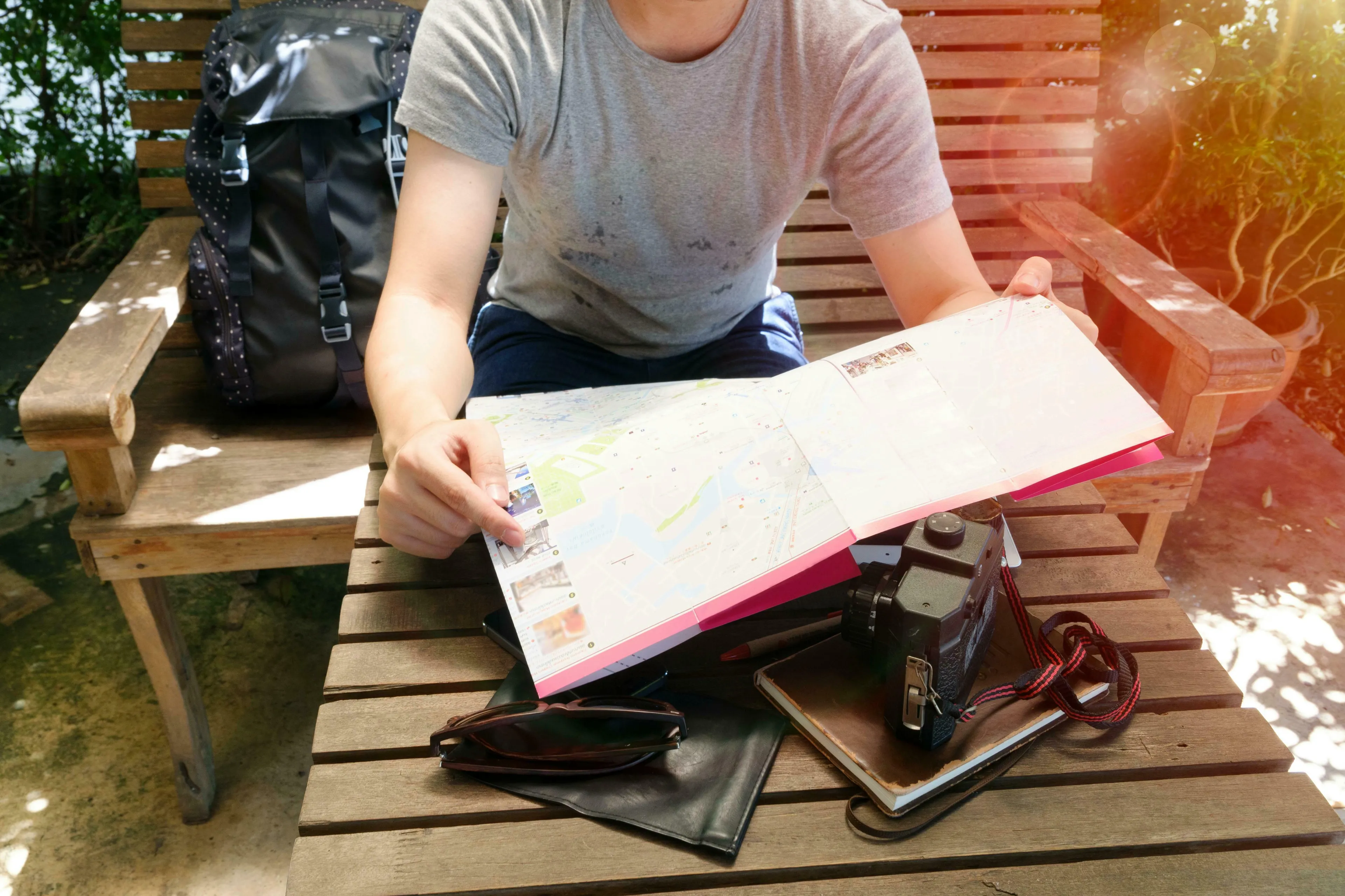A person sitting on a wooden bench, studying a map to plan a trip, with a backpack, camera, and sunglasses nearby.