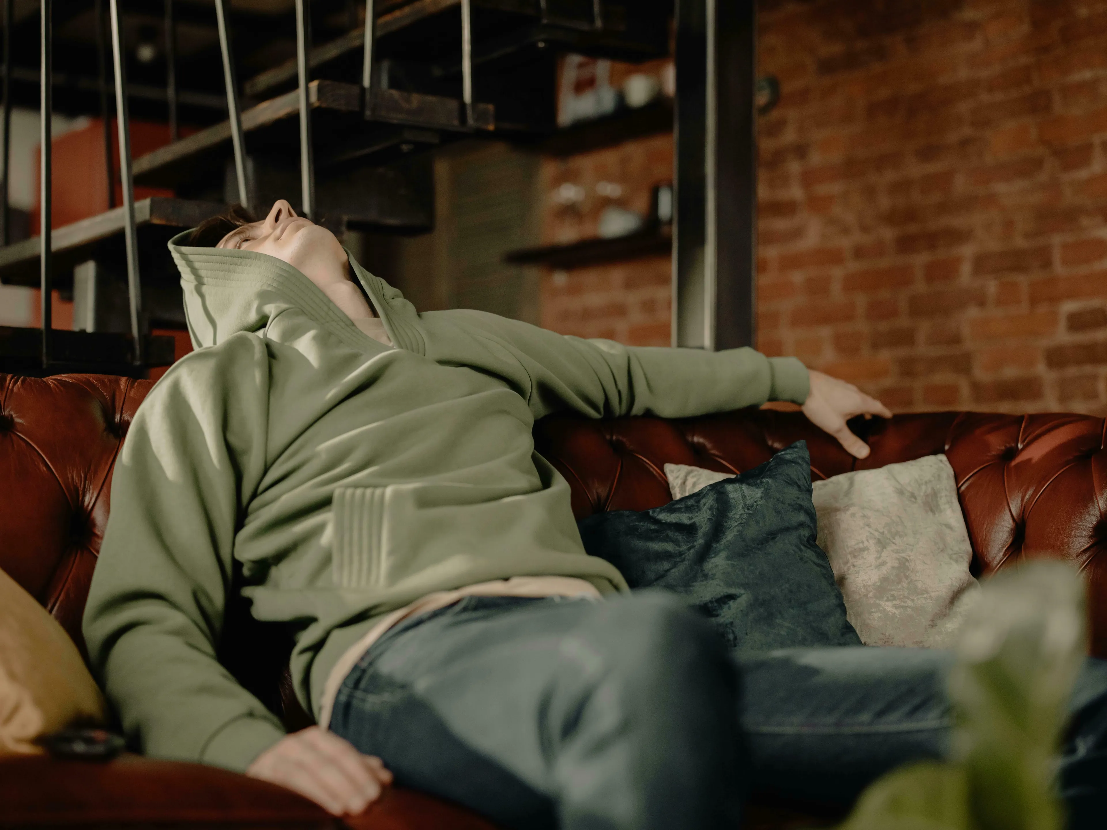 Man in a green hoodie lying on a brown leather couch, contemplating What's Your Procrastination Type or What do you do when you're bored.