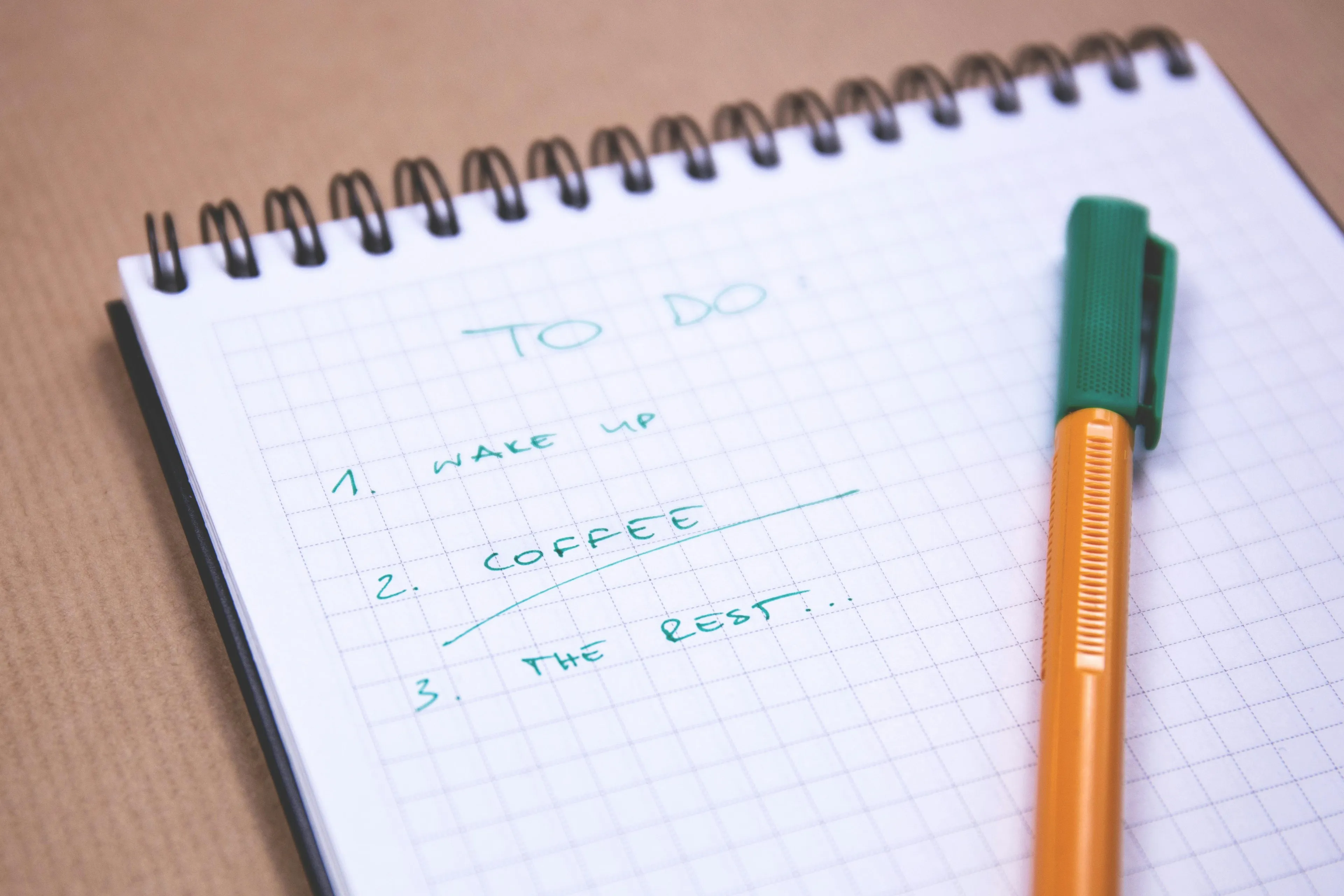 A grid-lined spiral notebook with a simple to-do list: Wake Up, Coffee, The Rest... A yellow pen lies on the right. Procrastination type.