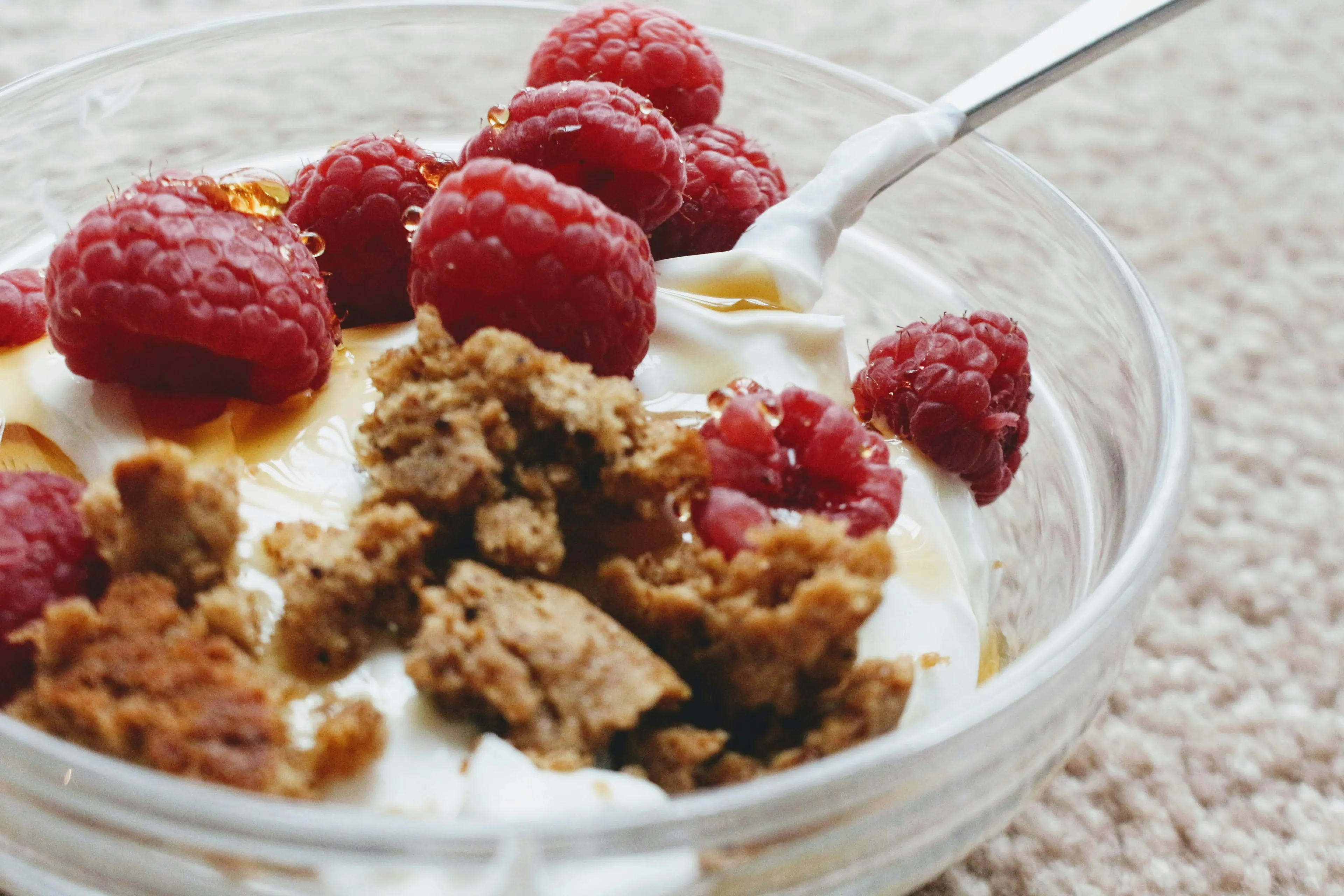 A close-up of delicious yogurt with toppings: fresh raspberries, granola, and honey drizzle in a clear bowl with a spoon.